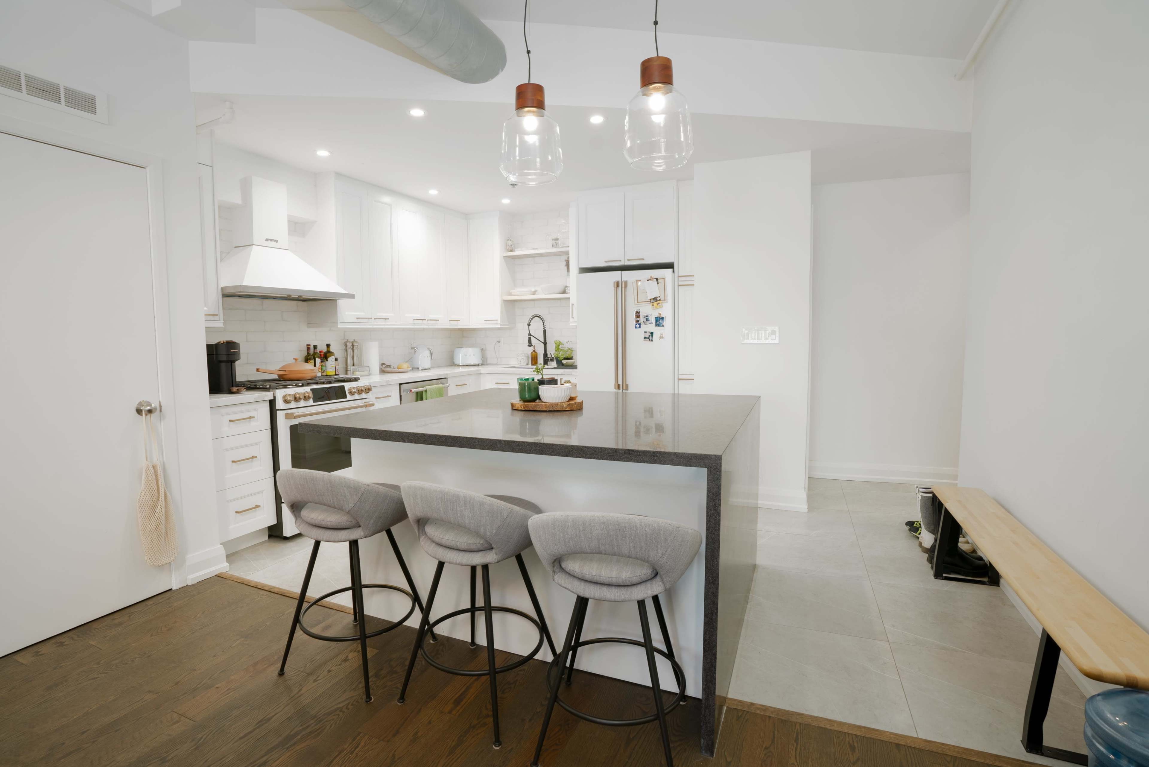 A modern kitchen featuring a central island with four bar stools, white cabinetry, stainless steel appliances, and a mix of tile and hardwood flooring.