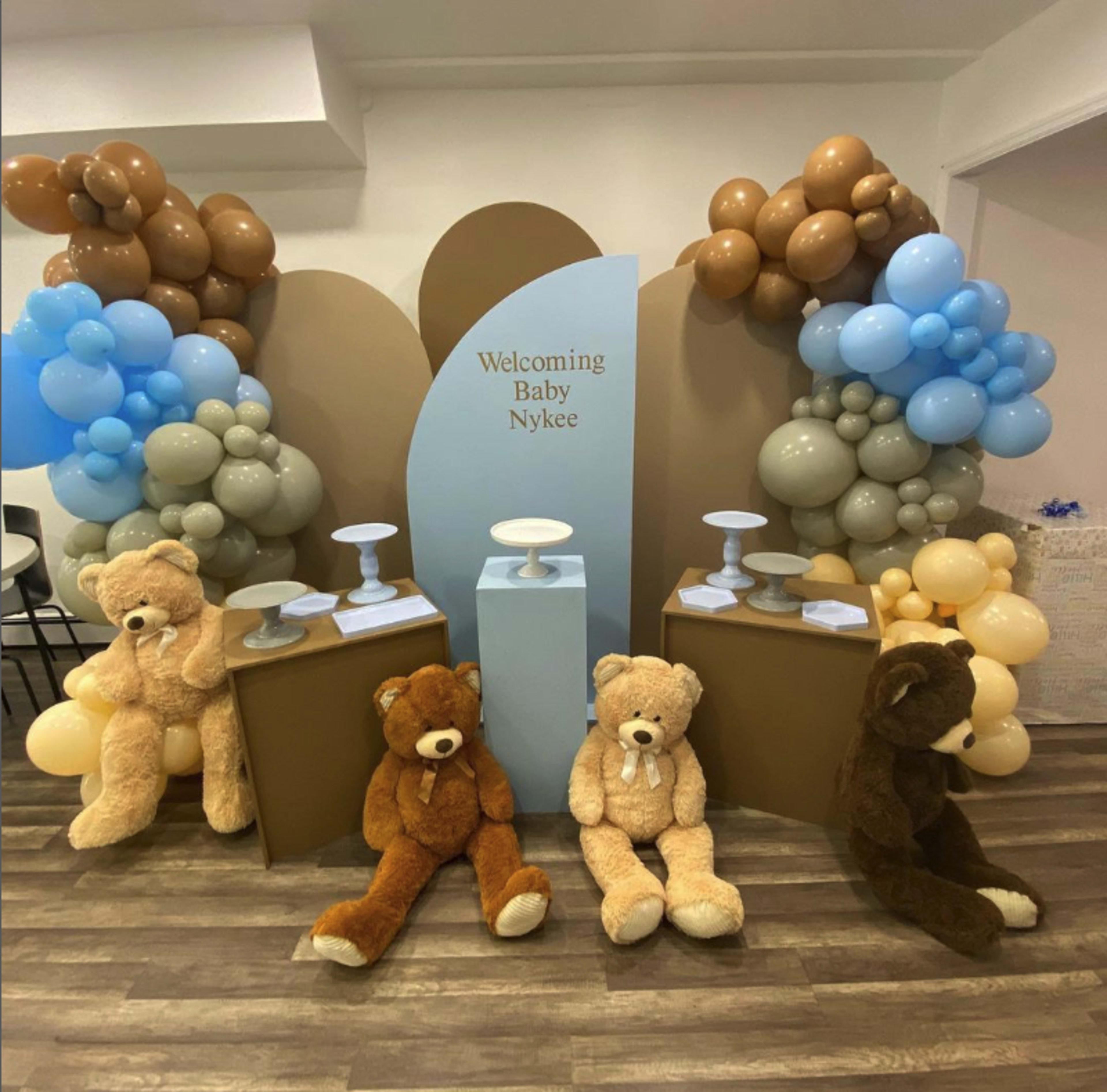 The scene features a decorated area for a baby welcoming event, with a backdrop of balloons in shades of blue, brown, and gray, flanked by large teddy bears sitting on the floor.