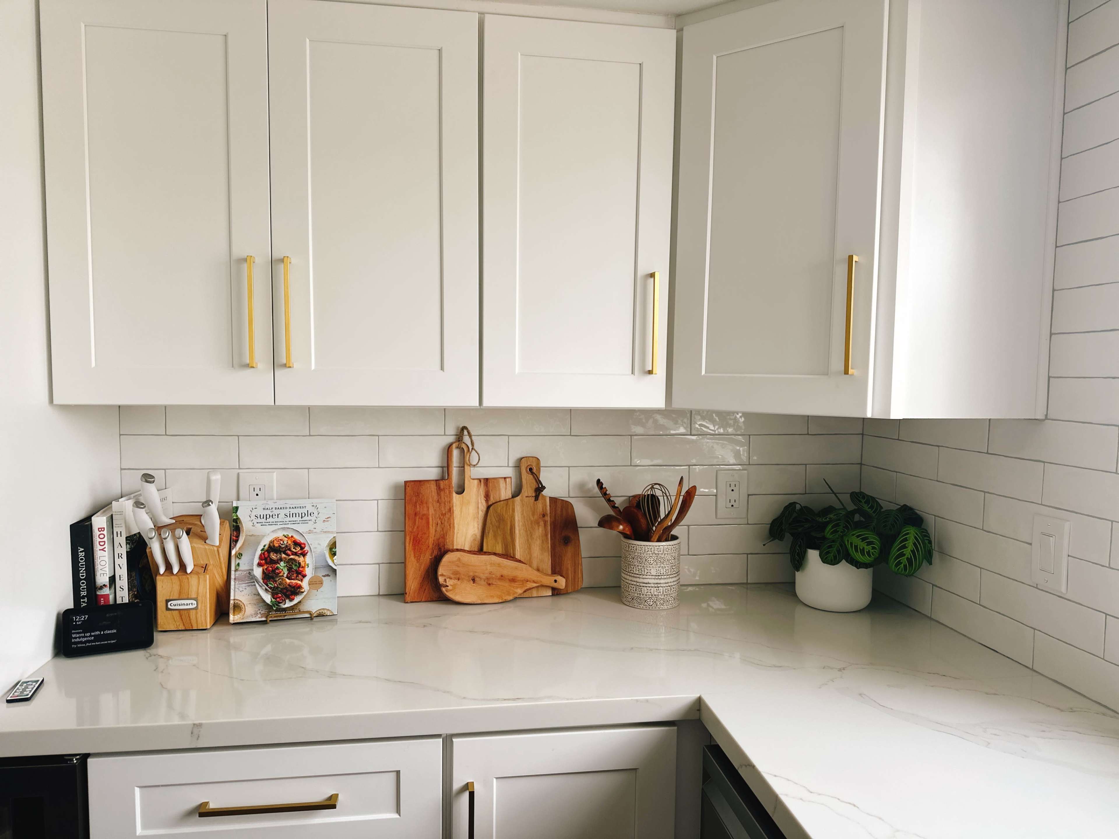 A clean kitchen countertop features white cabinetry, cutting boards, utensils in a container, a plant, and a recipe book.