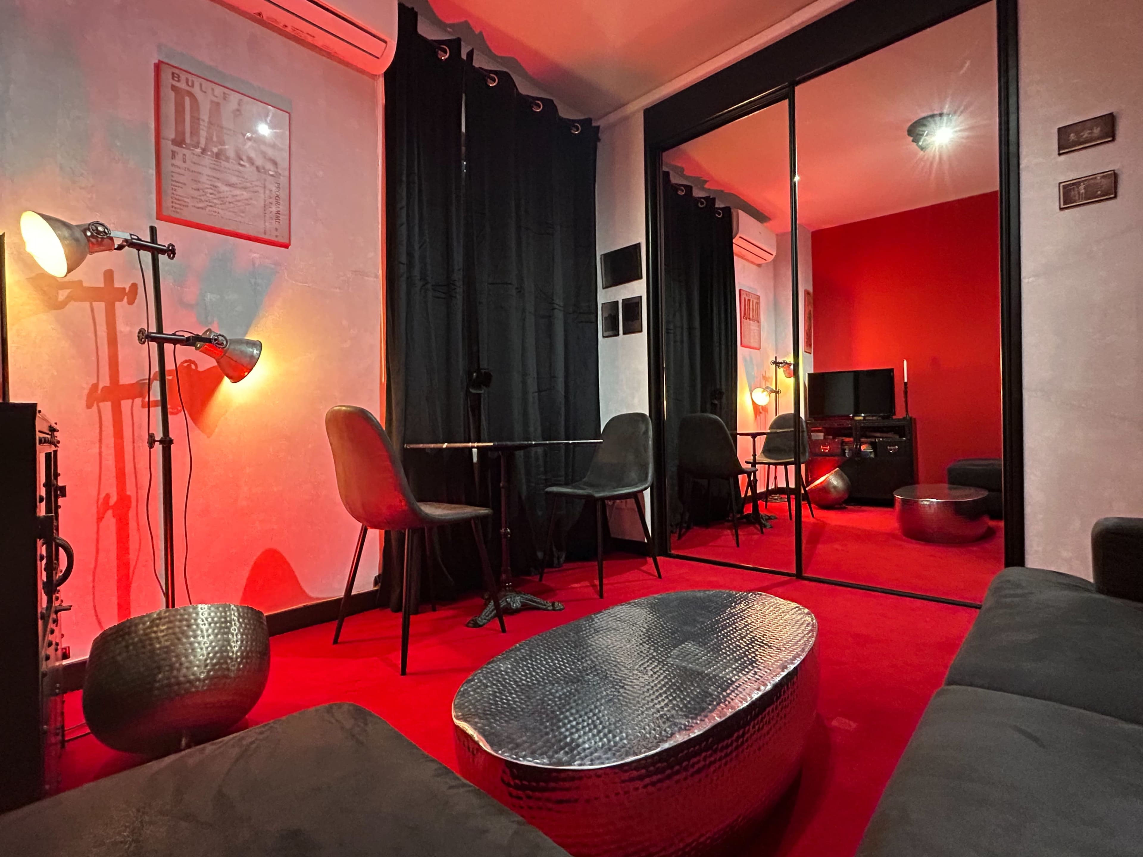 The image depicts a lounge area with red walls, black curtains, and modern furniture, including a mirrored coffee table and several chairs around a small table.
