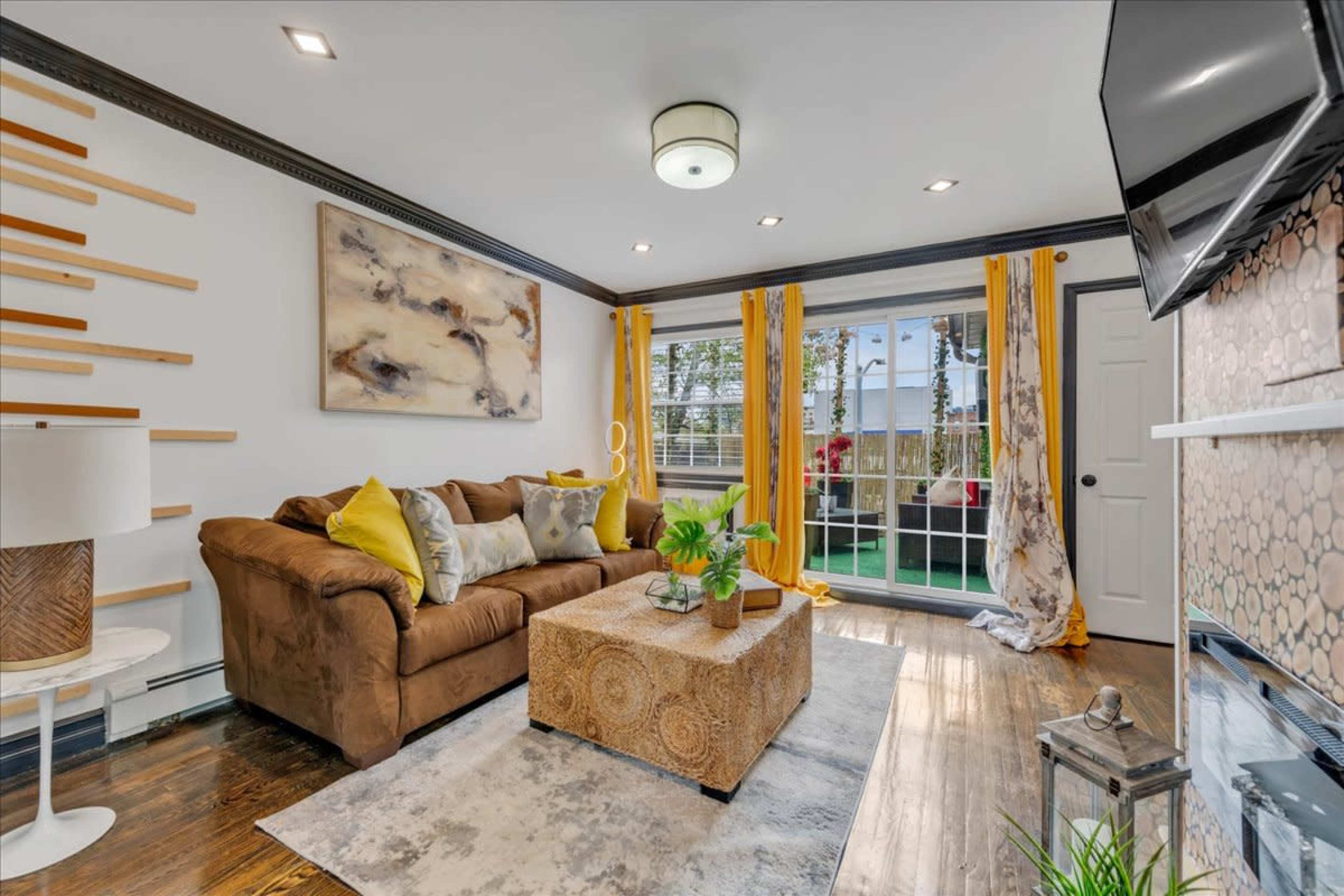 The living room features a brown couch with colorful pillows, a patterned coffee table, and large windows dressed with yellow curtains.
