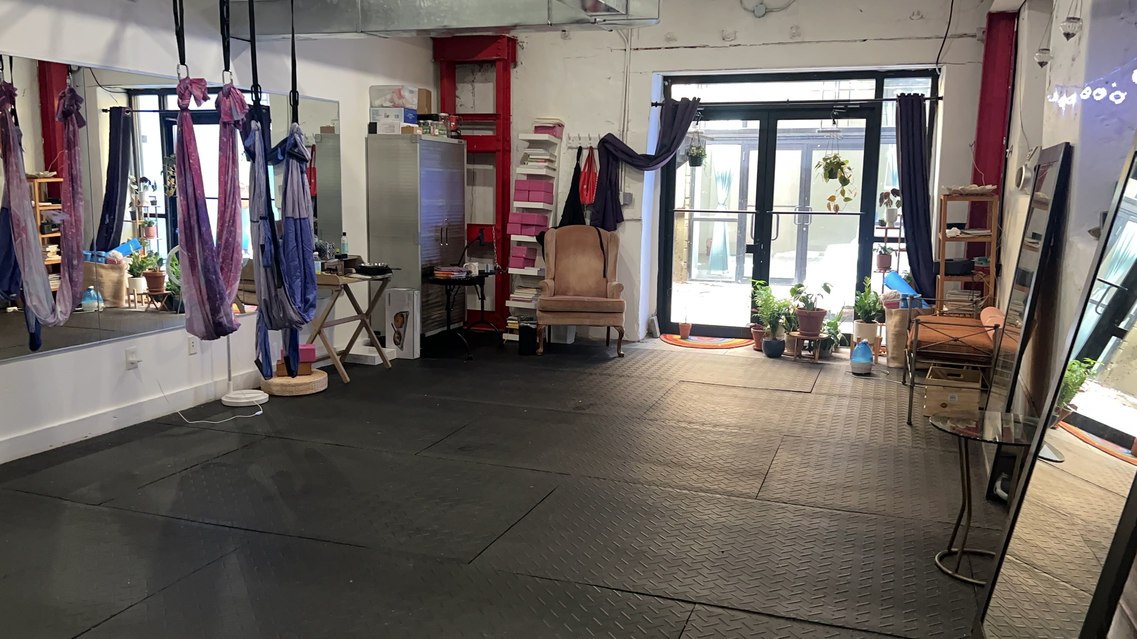 An indoor space with exercise mats, aerial silks hanging from the ceiling, and a mix of furniture and plants arranged against the walls.