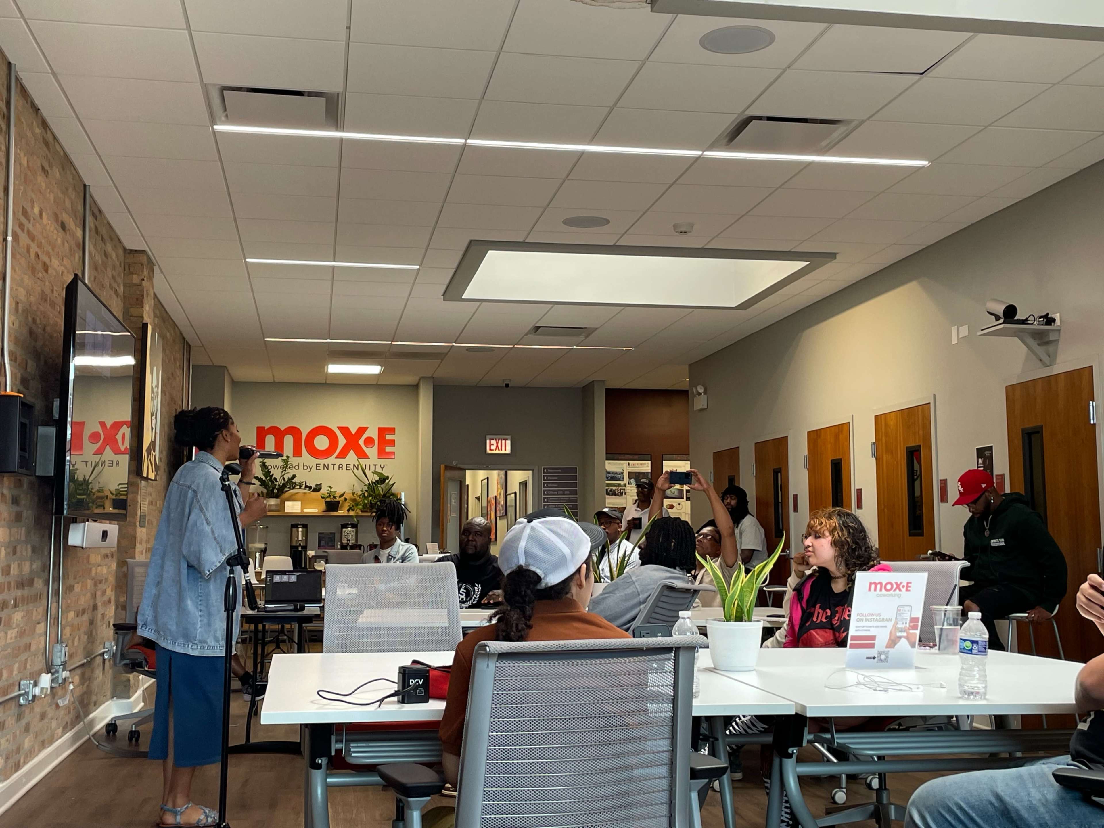 A woman stands in front of an audience in a modern room with brick walls, engaging them during a presentation or meeting.