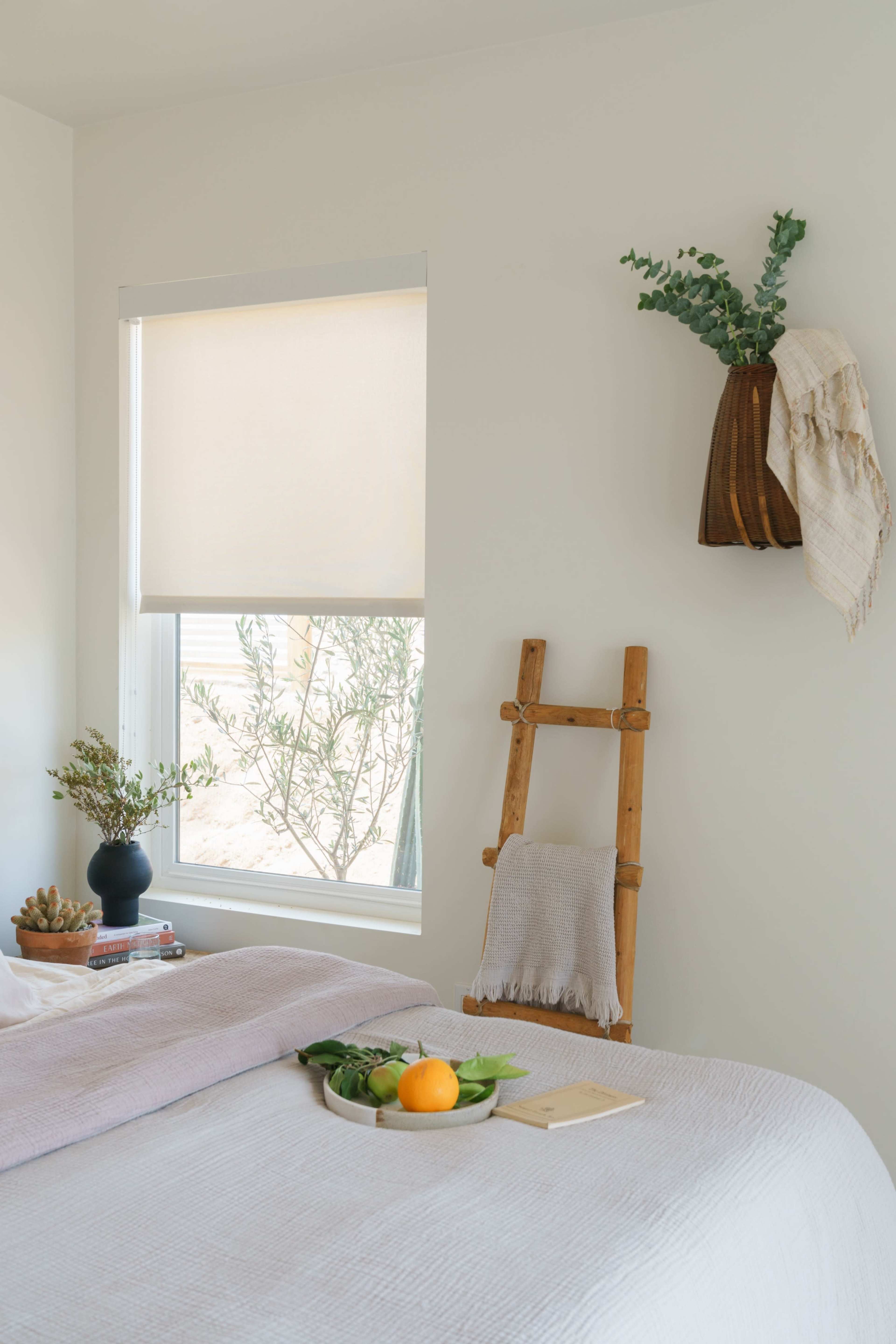 A simple bedroom features a bed with a light-colored blanket, a wooden ladder with a blanket, a small black vase with greenery, and a window with a roller shade.