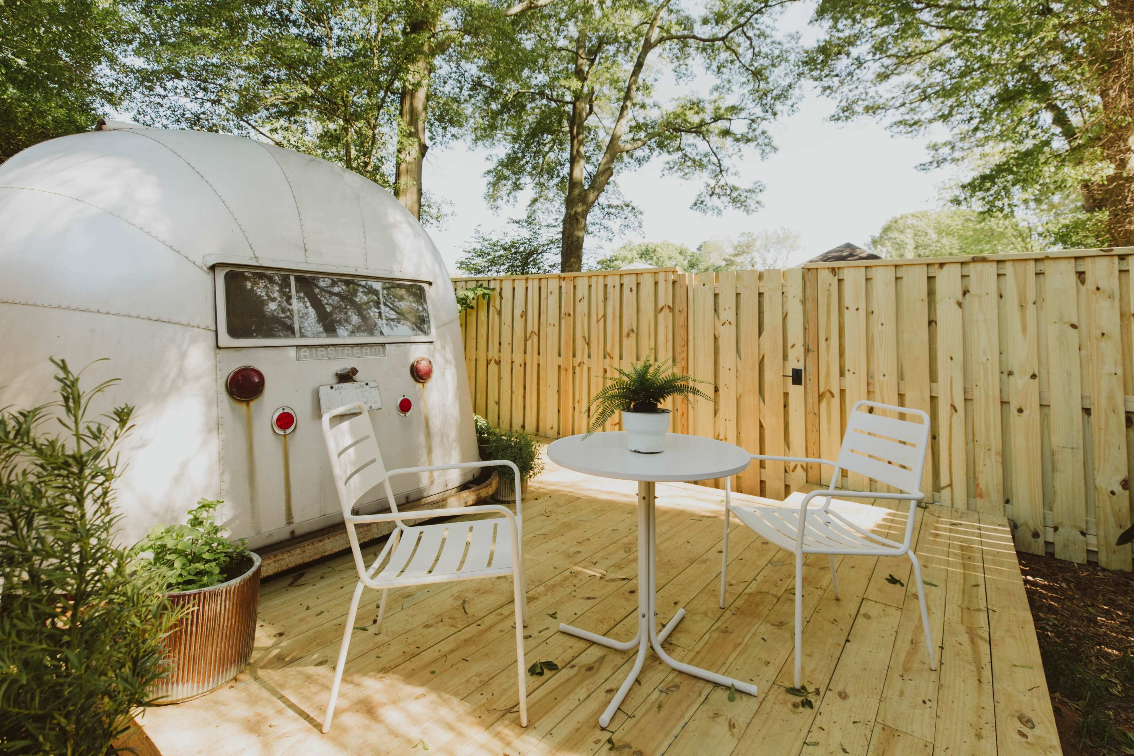 A vintage trailer is parked beside a small wooden deck with two white chairs and a table, surrounded by a wooden fence and greenery.