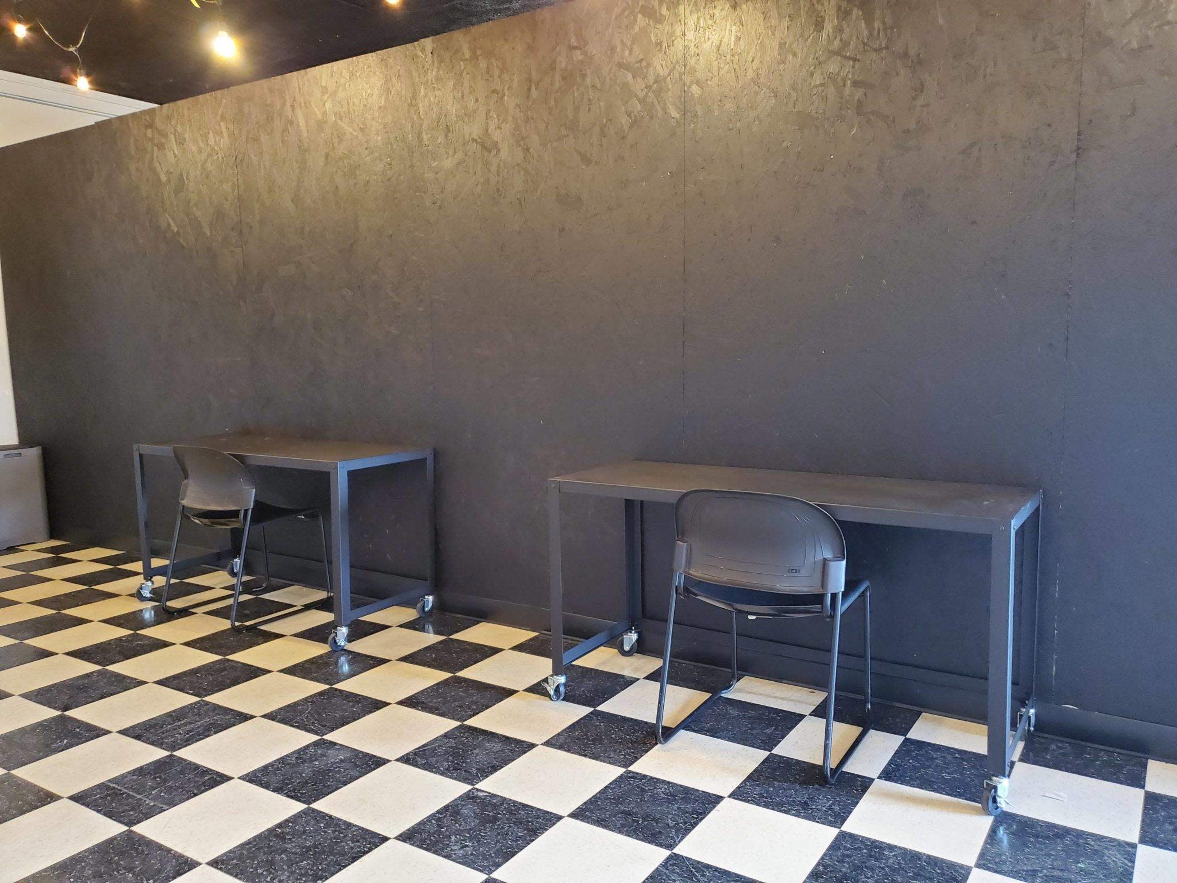 Two mobile black tables with rolling chairs are positioned against a black wall in a room with a checkerboard floor pattern.