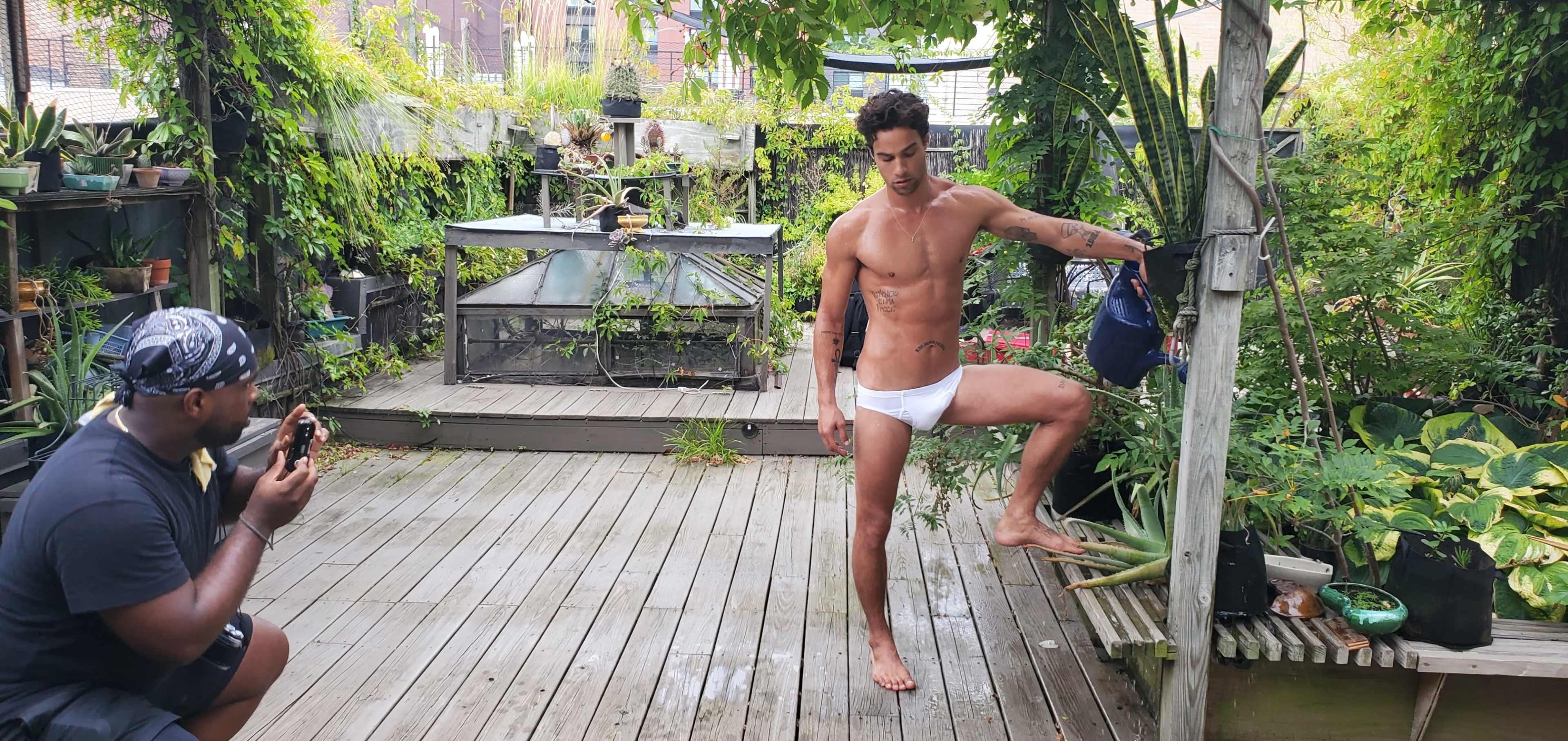 A man in white swimwear poses on a wooden deck surrounded by greenery while another man takes a photo of him.