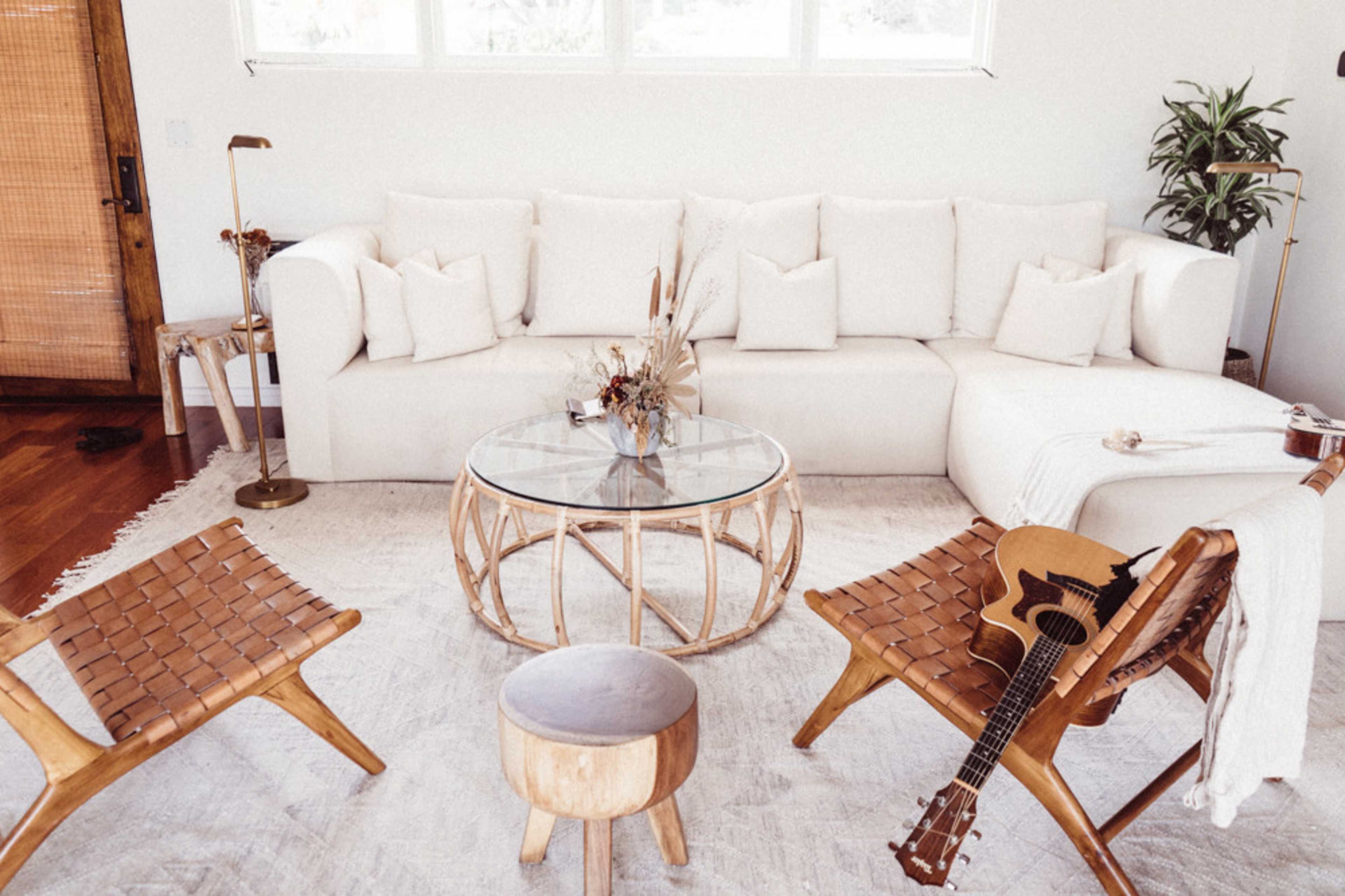 A minimalist living room with a large white sofa, a round glass coffee table, two wooden chairs, a small wooden stool, and a guitar arranged on a light-colored rug.
