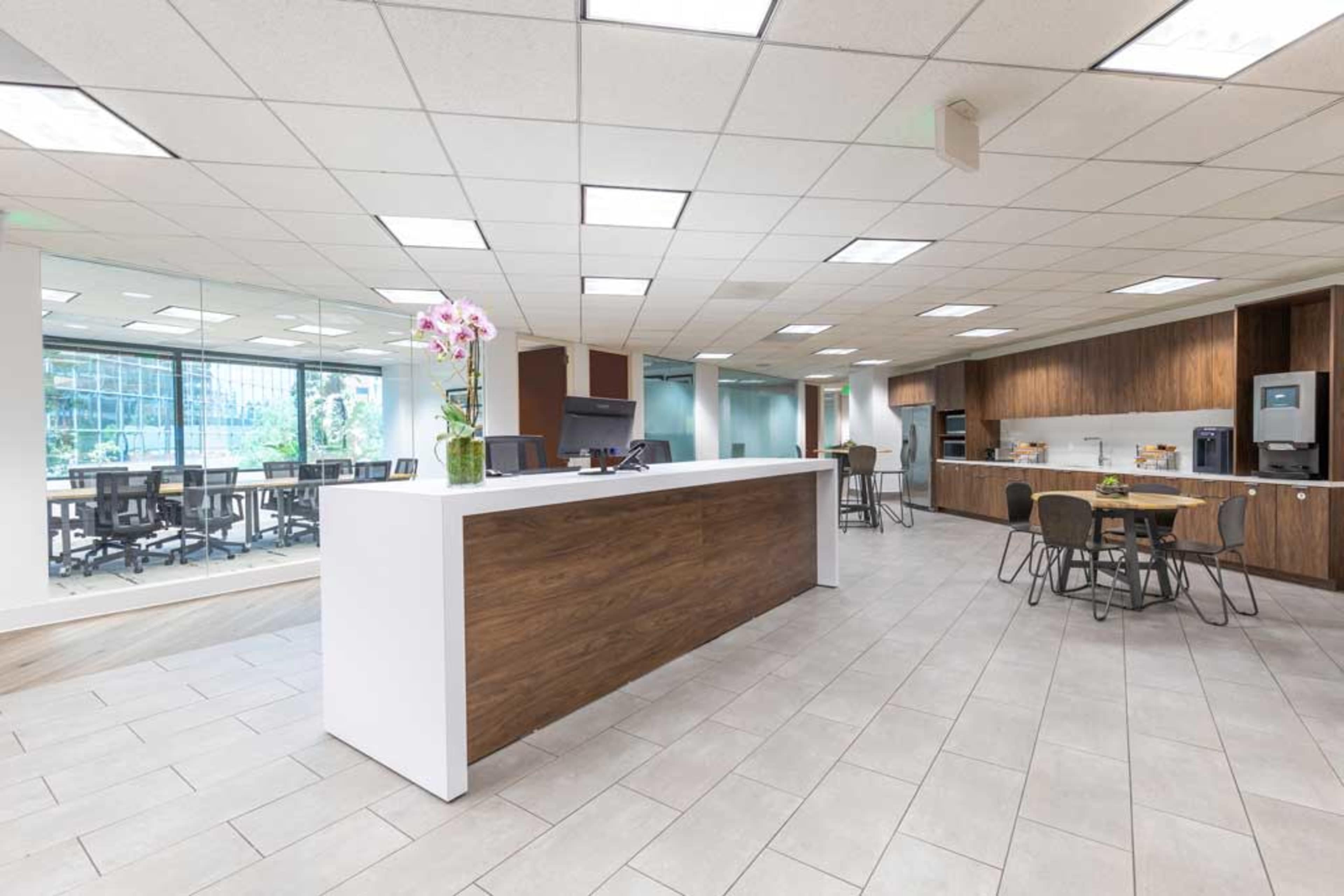 A modern office break room with a reception area, a dining space with tables and chairs, and a kitchenette featuring appliances.