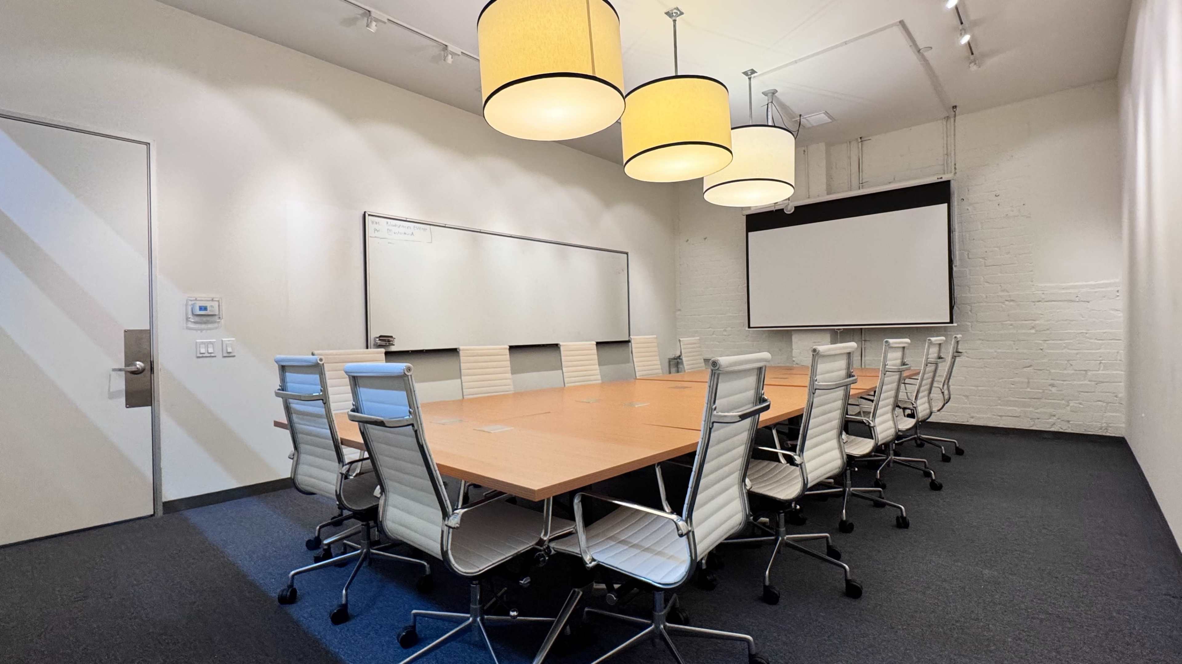 A large conference room features a long wooden table surrounded by ergonomic rolling chairs, with pendant lighting overhead and a projector screen on the wall.