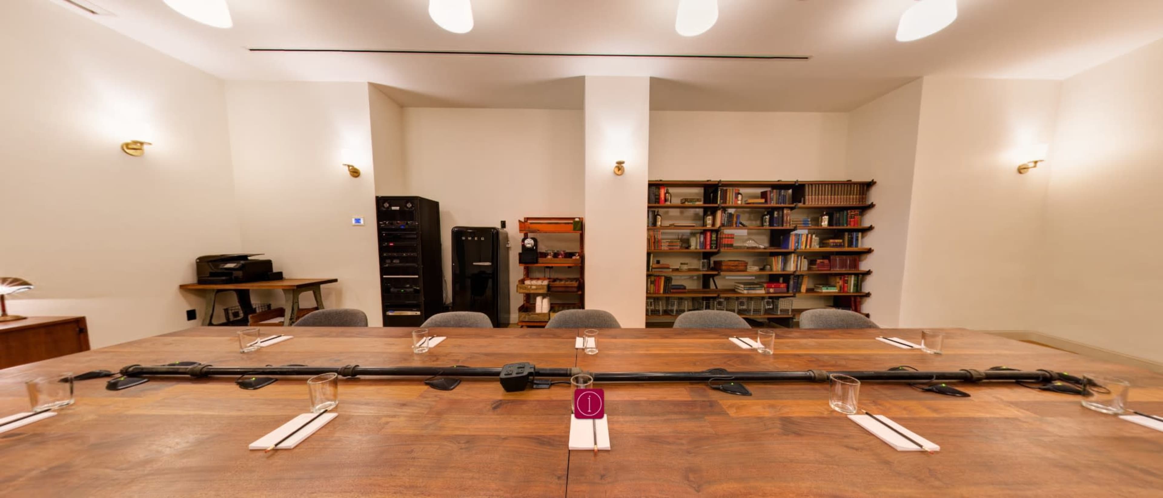 A large wooden table is set for a meeting, featuring water glasses and utensils, with a book-filled shelf and a black appliance in the background.