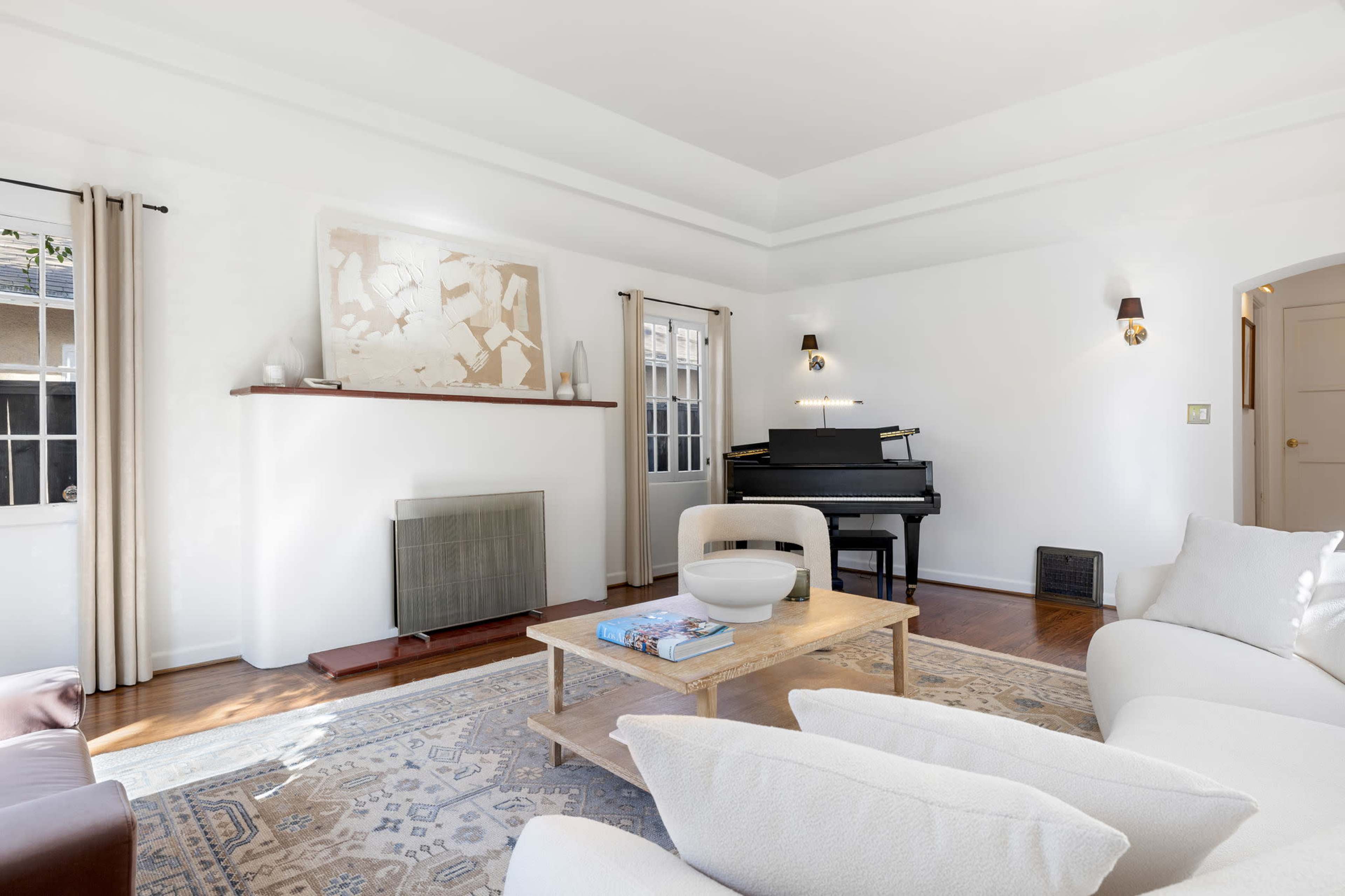 A light-filled living room features a piano, a coffee table, and a patterned area rug, with large windows and minimalistic decor.
