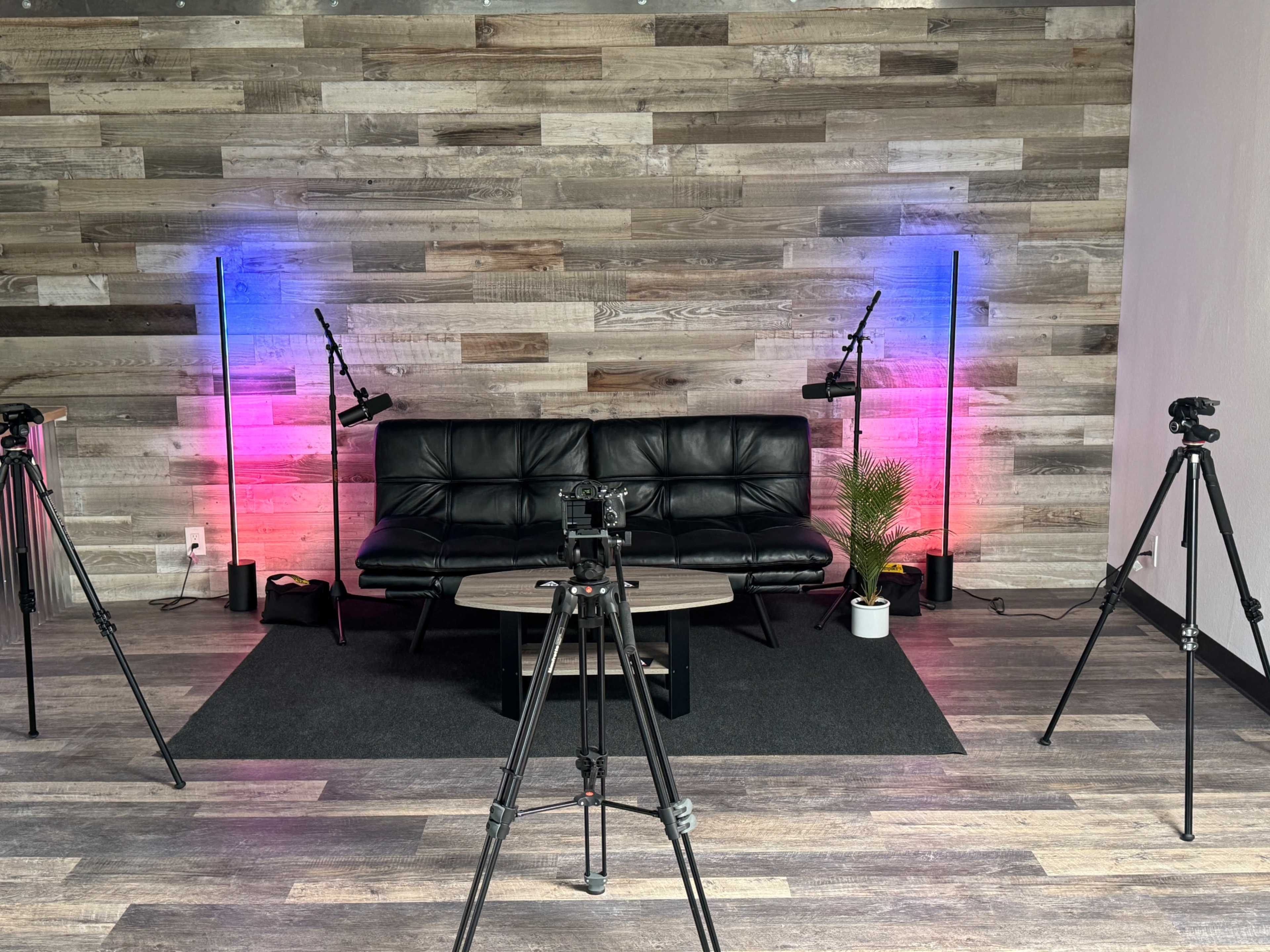A modern studio setup with a black couch, a round table, and several tripods positioned around the space, illuminated by colorful lights on the walls.