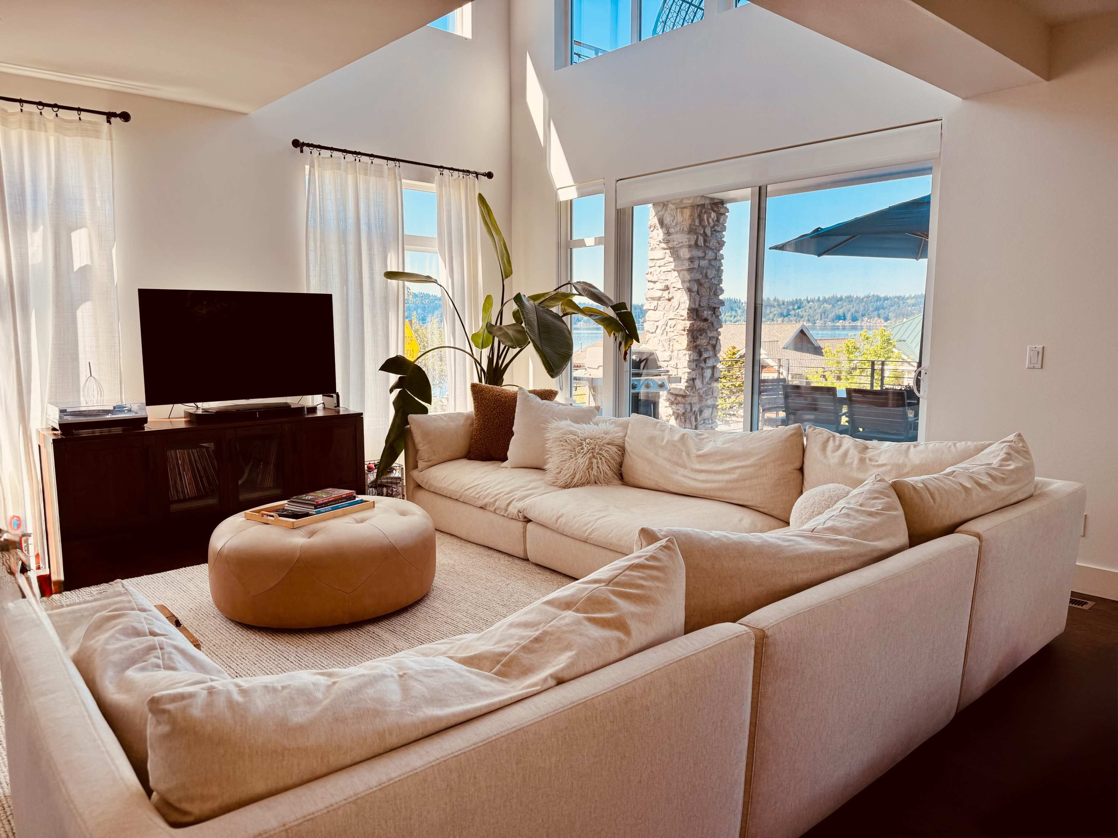 The living room features a sectional sofa, an ottoman, and large windows that offer a view of the outdoors.