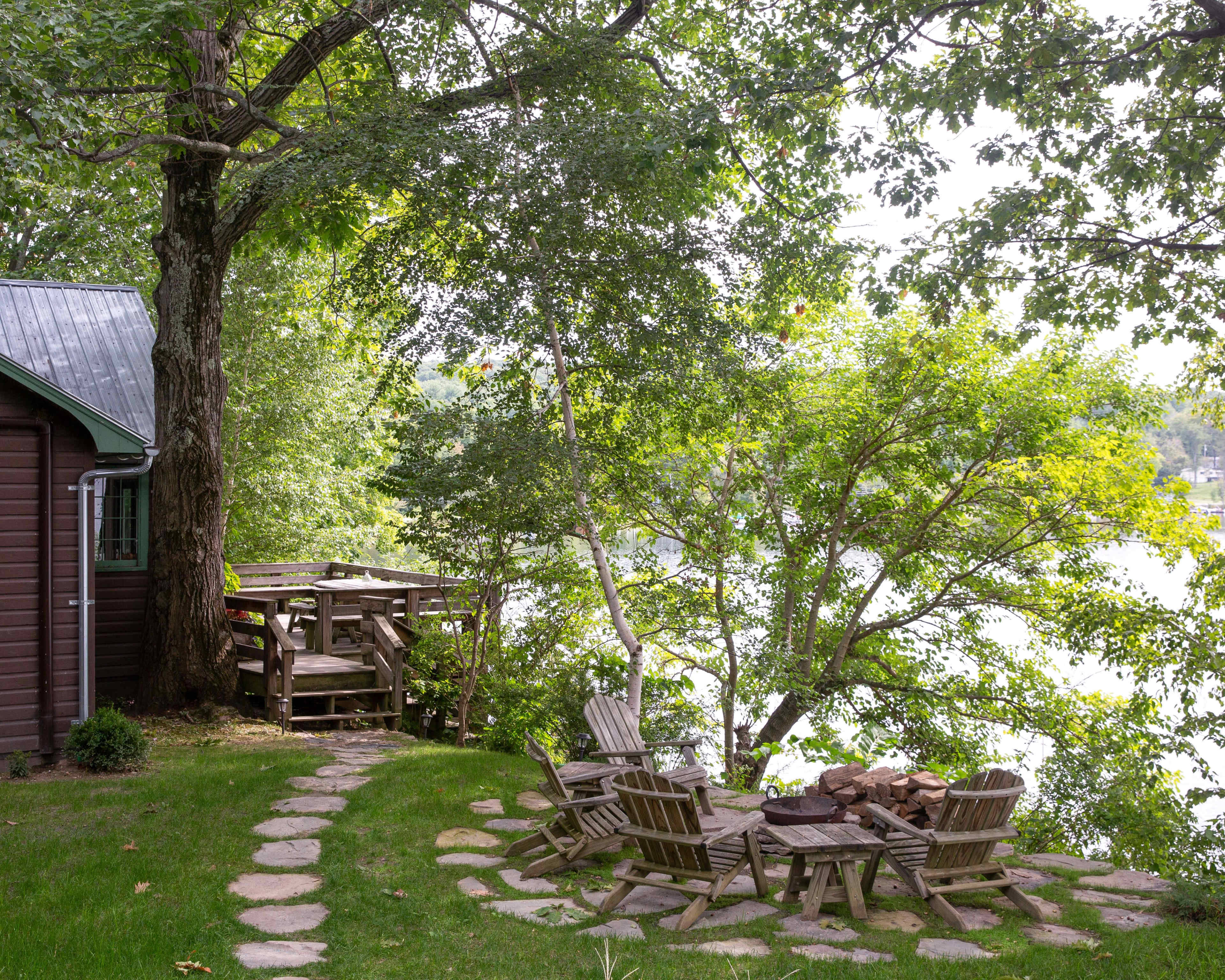 A circular stone patio with wooden chairs surrounds a fire pit, situated near a house by a tree-lined water's edge.