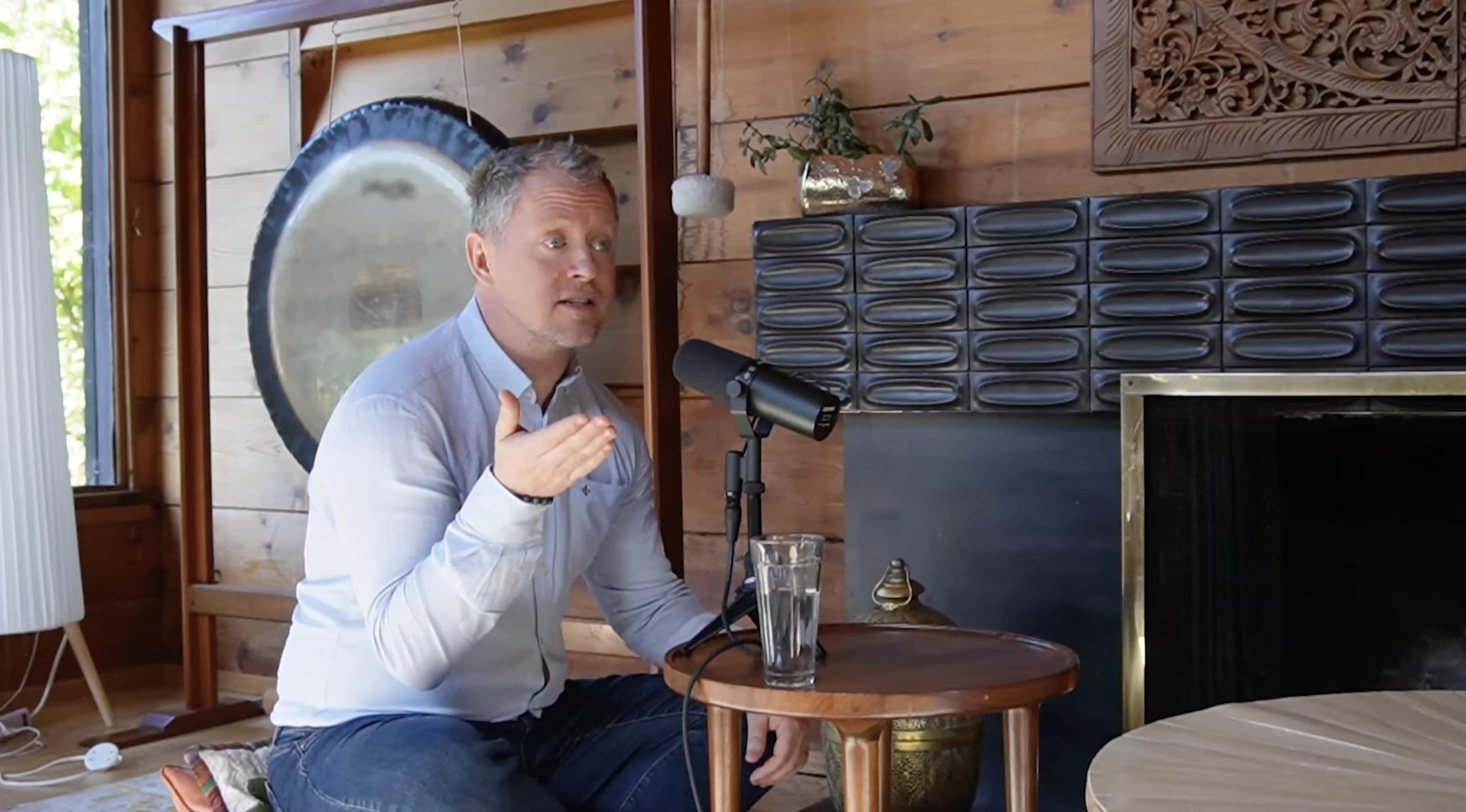 A man is sitting at a wooden table with a microphone in front of him, gesturing as he speaks, in a room adorned with wooden paneling and decorative items.