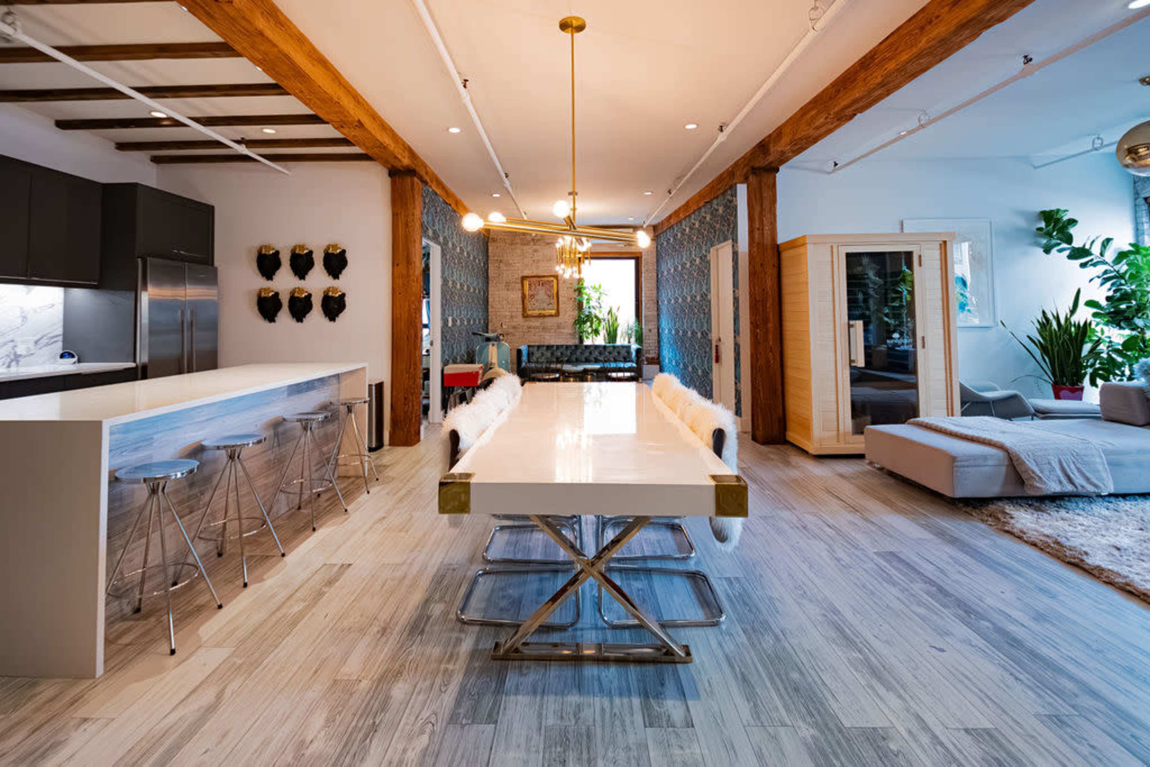 The image shows a spacious modern dining area with a large white table, sleek metal chairs, exposed wooden beams, and decorative plants throughout the room.