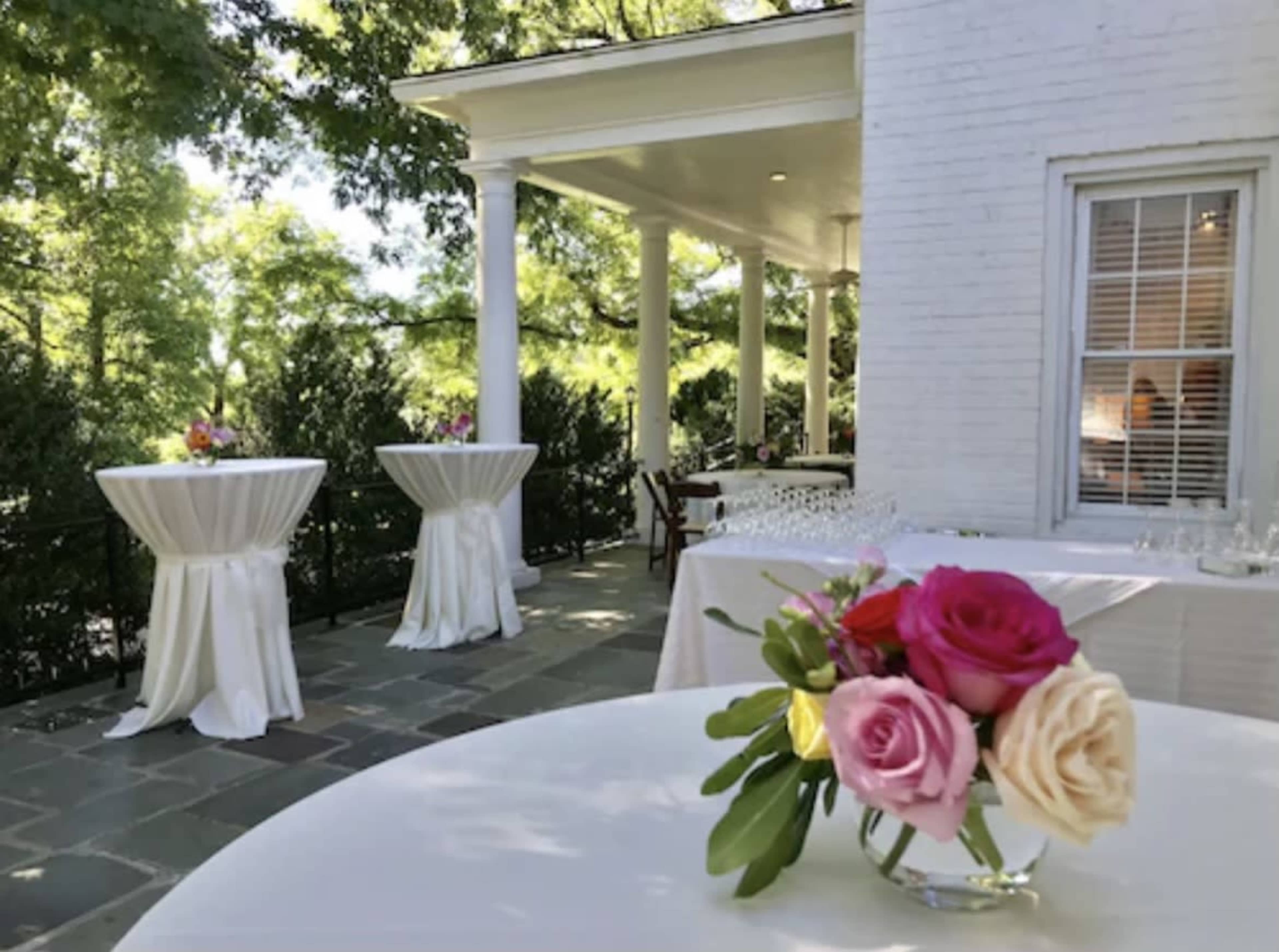 The image shows a outdoor venue with white tablecloths on round tables, adorned with floral centerpieces, set against a backdrop of greenery and a white building.