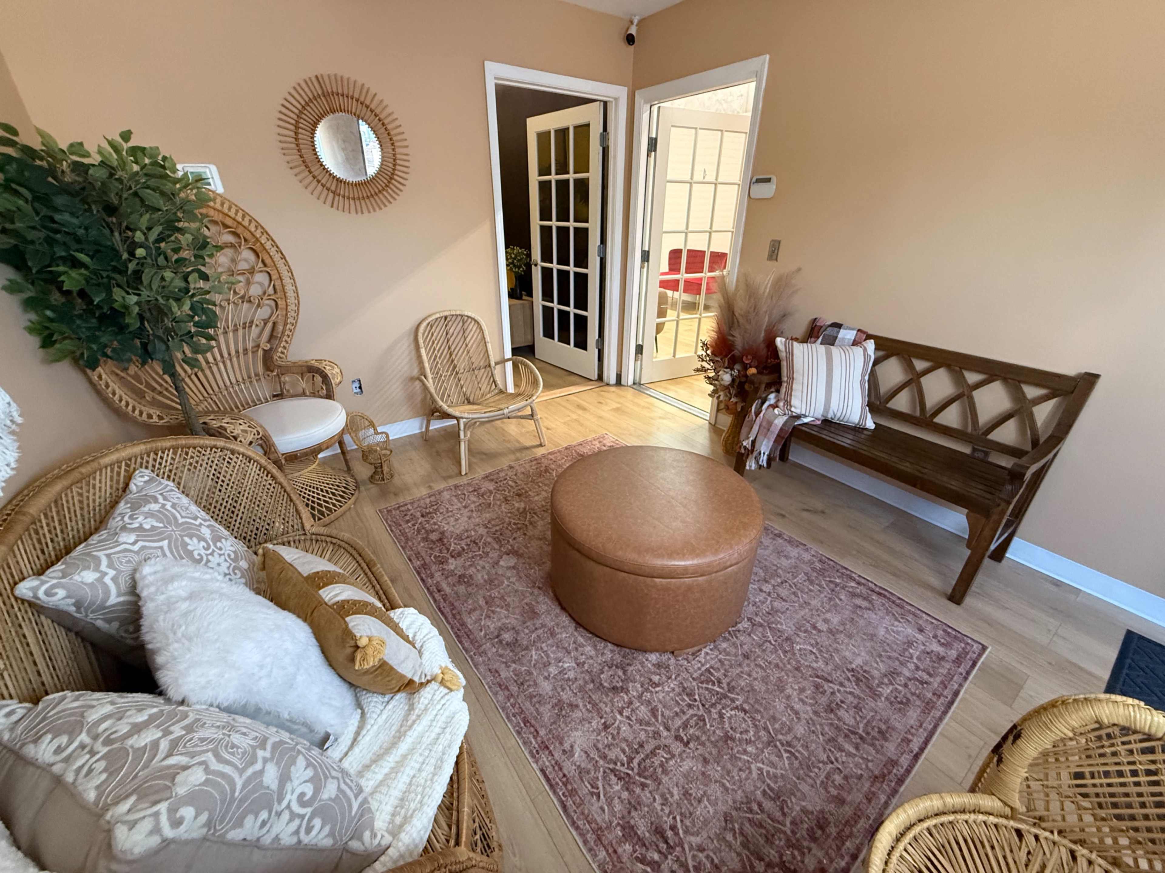 The image shows a cozy living area with wicker furniture, a round leather ottoman, and a doorway leading to another room.