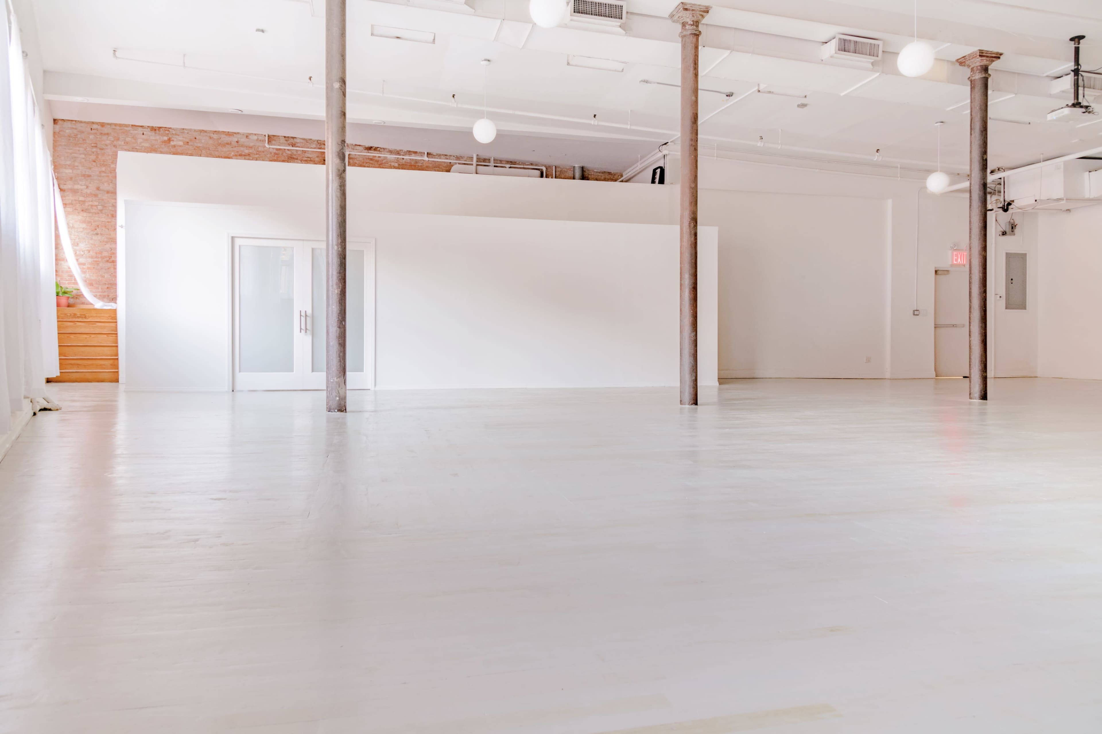 The image shows a spacious, empty studio with white walls, wooden pillars, and large windows allowing natural light.
