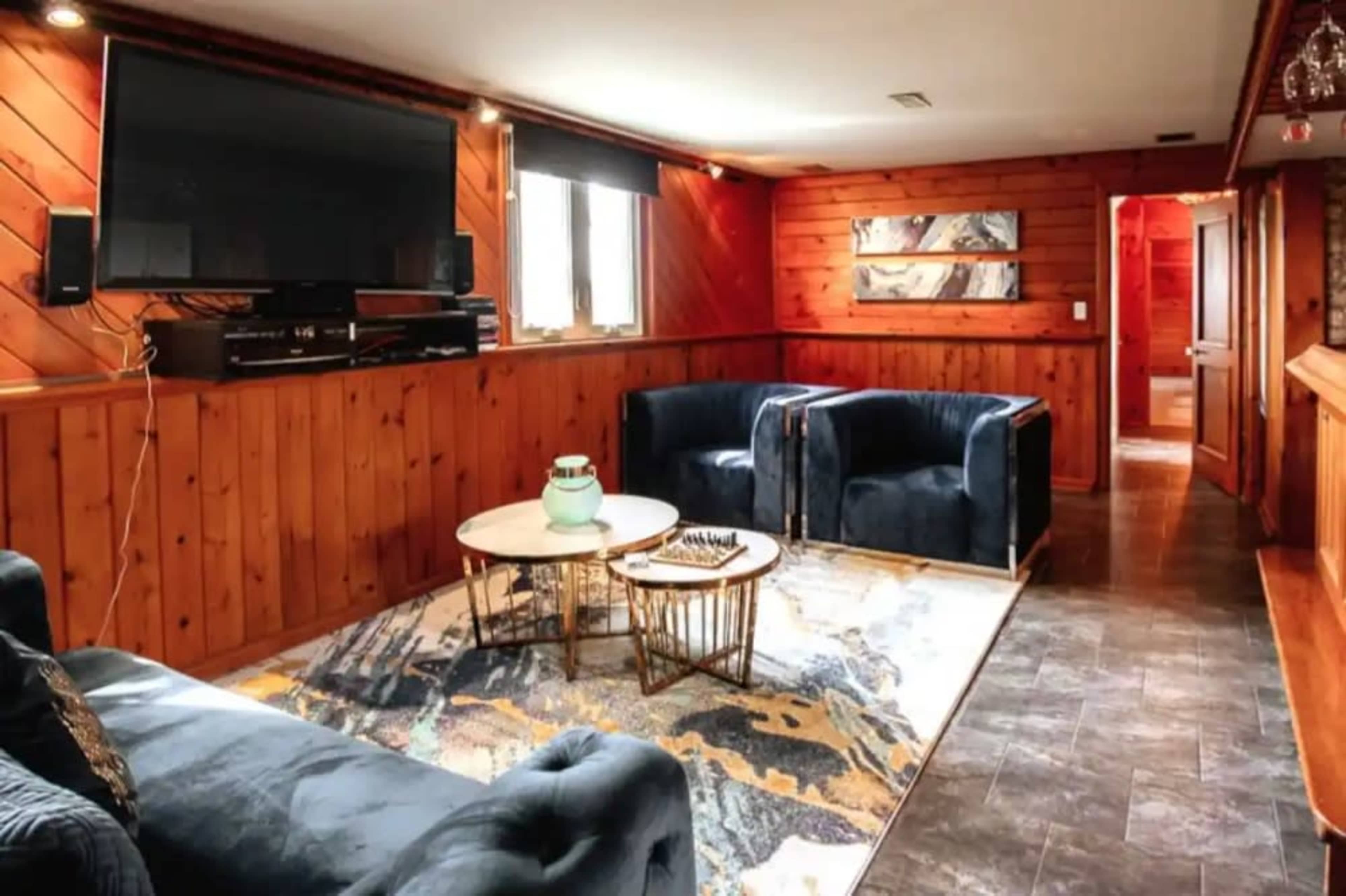 A cozy basement lounge area with wooden walls, two blue armchairs, a coffee table, and a large television mounted on the wall.
