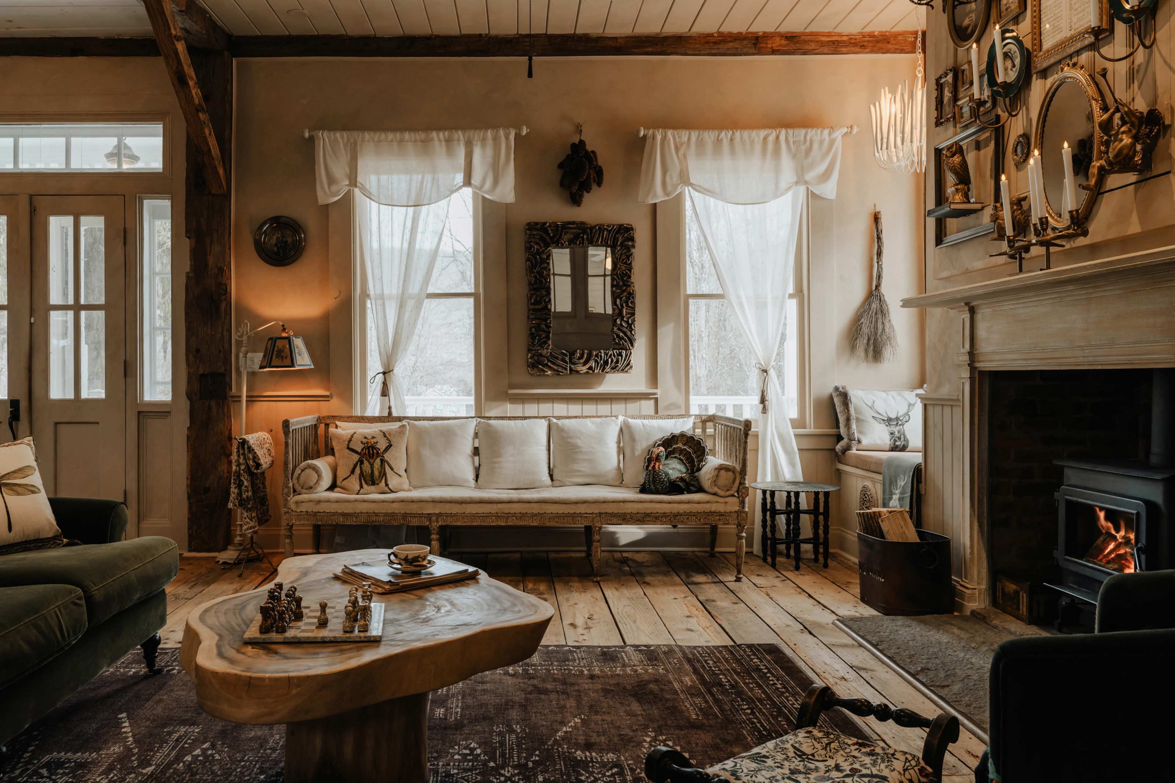The image shows a cozy living room with large windows, a wooden floor, a fireplace, and a sofa adorned with cushions.