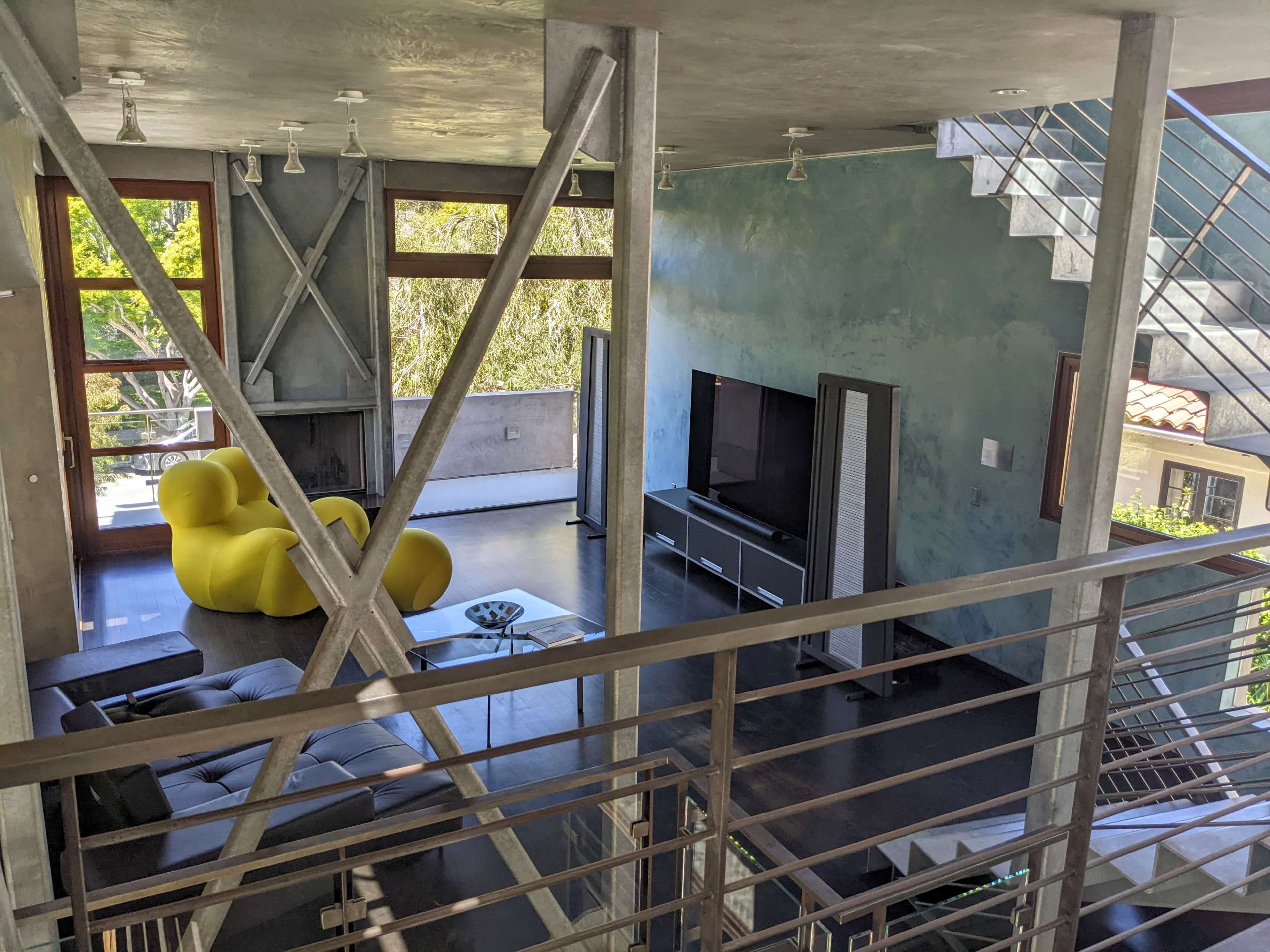 The image shows a modern, two-story living area with a yellow chair, a gray sofa, and large windows allowing natural light to fill the space.