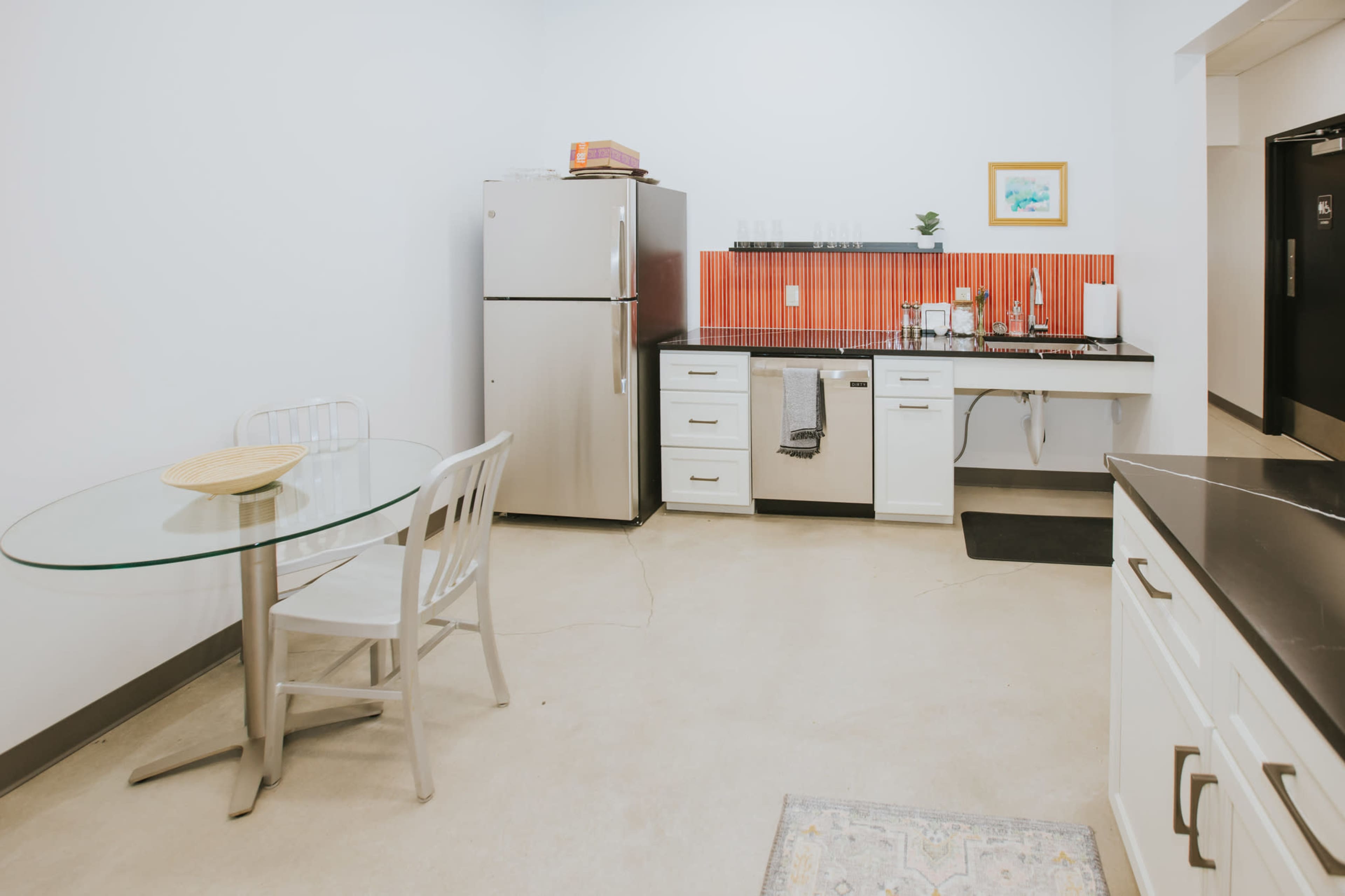 The image shows a modern kitchen with a round glass dining table, stainless steel refrigerator, and a countertop with a sink and various kitchen items.