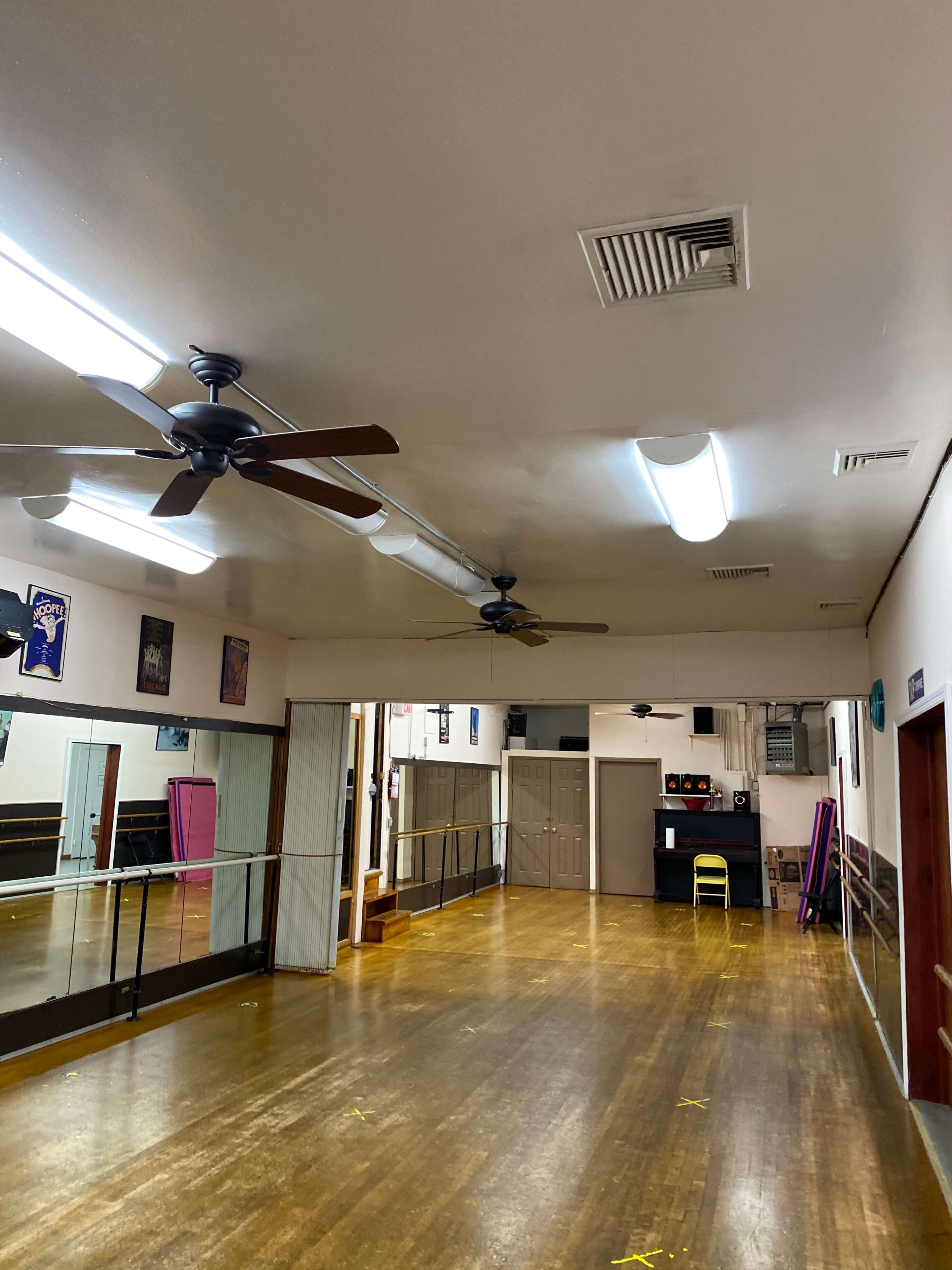 A dance studio with wooden flooring, wall-mounted mirrors, and two ceiling fans.