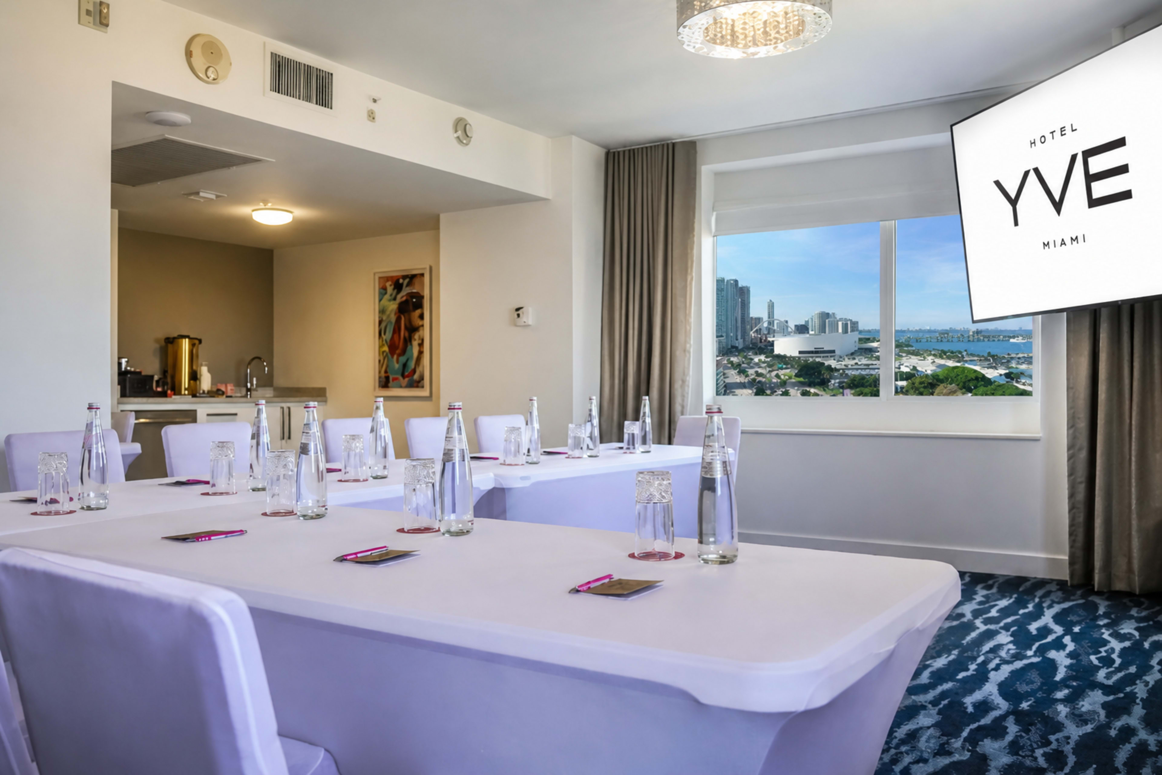 A conference room at Hotel YVE Miami features several tables set with water bottles and a presentation screen displaying the hotel’s logo, with a view of the city skyline through a window.