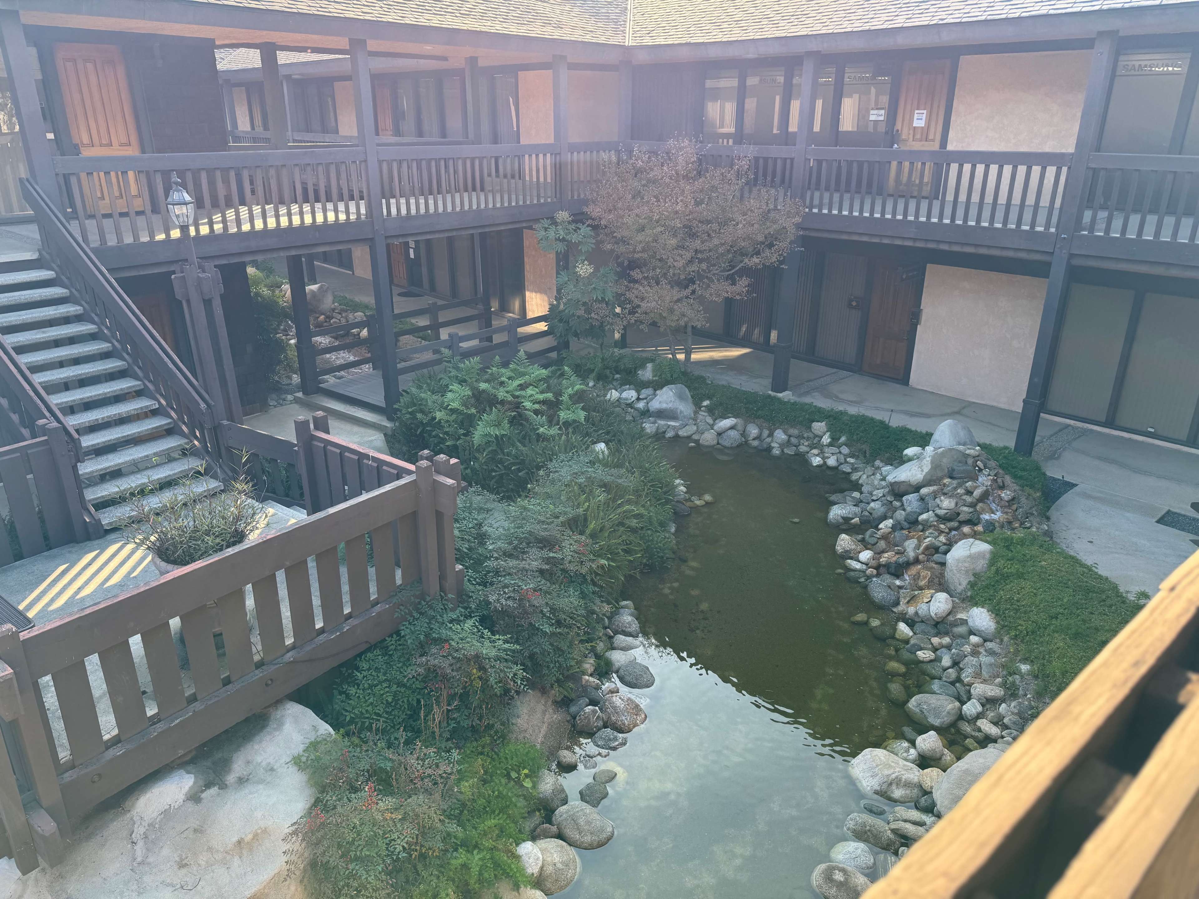 A tranquil courtyard with a pond surrounded by rocks and greenery, framed by wooden walkways and buildings in a multi-level structure.