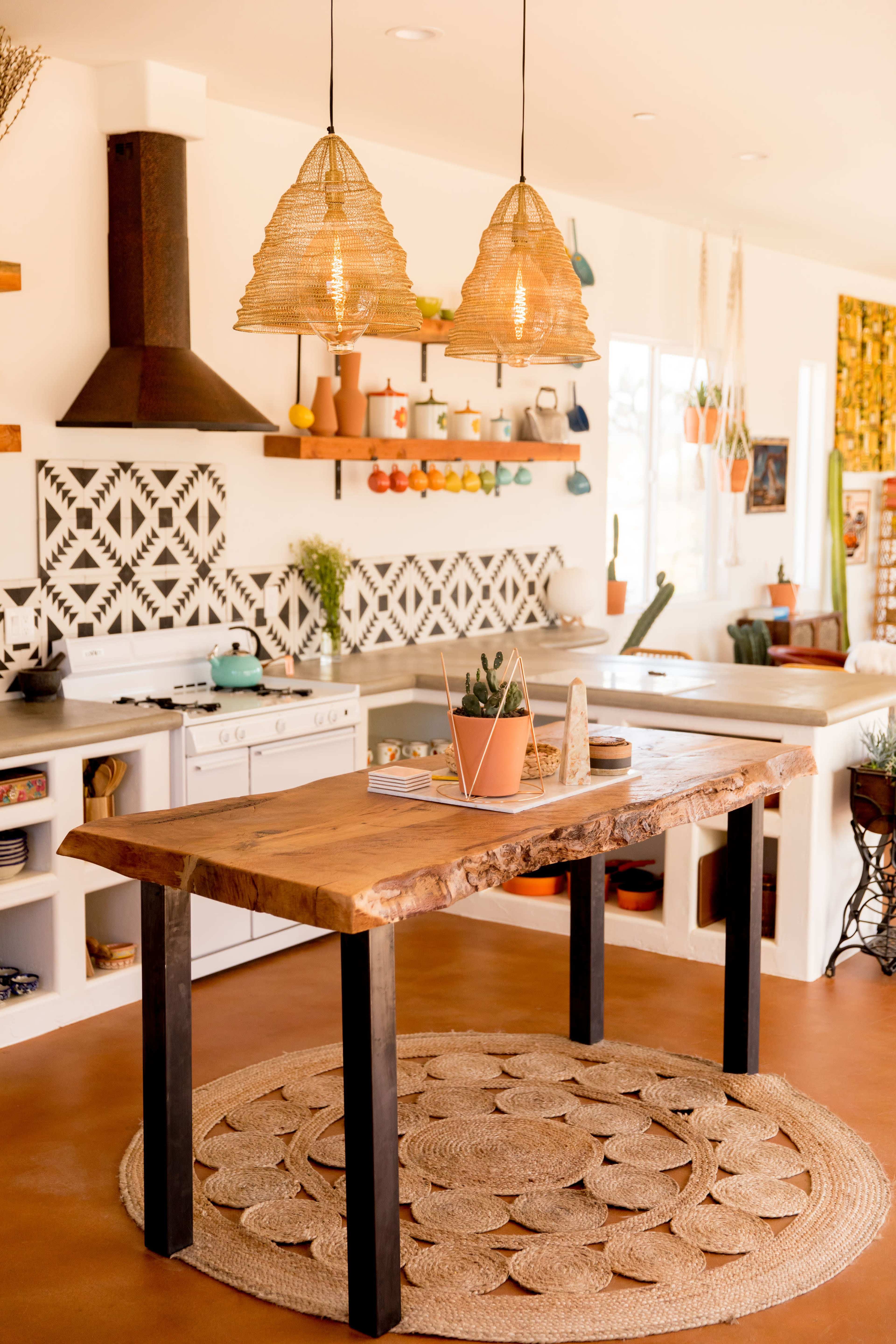 A modern kitchen features a wooden table with black legs, surrounded by a circular rug, under woven pendant lights, and decorated with colorful dishes on shelves.