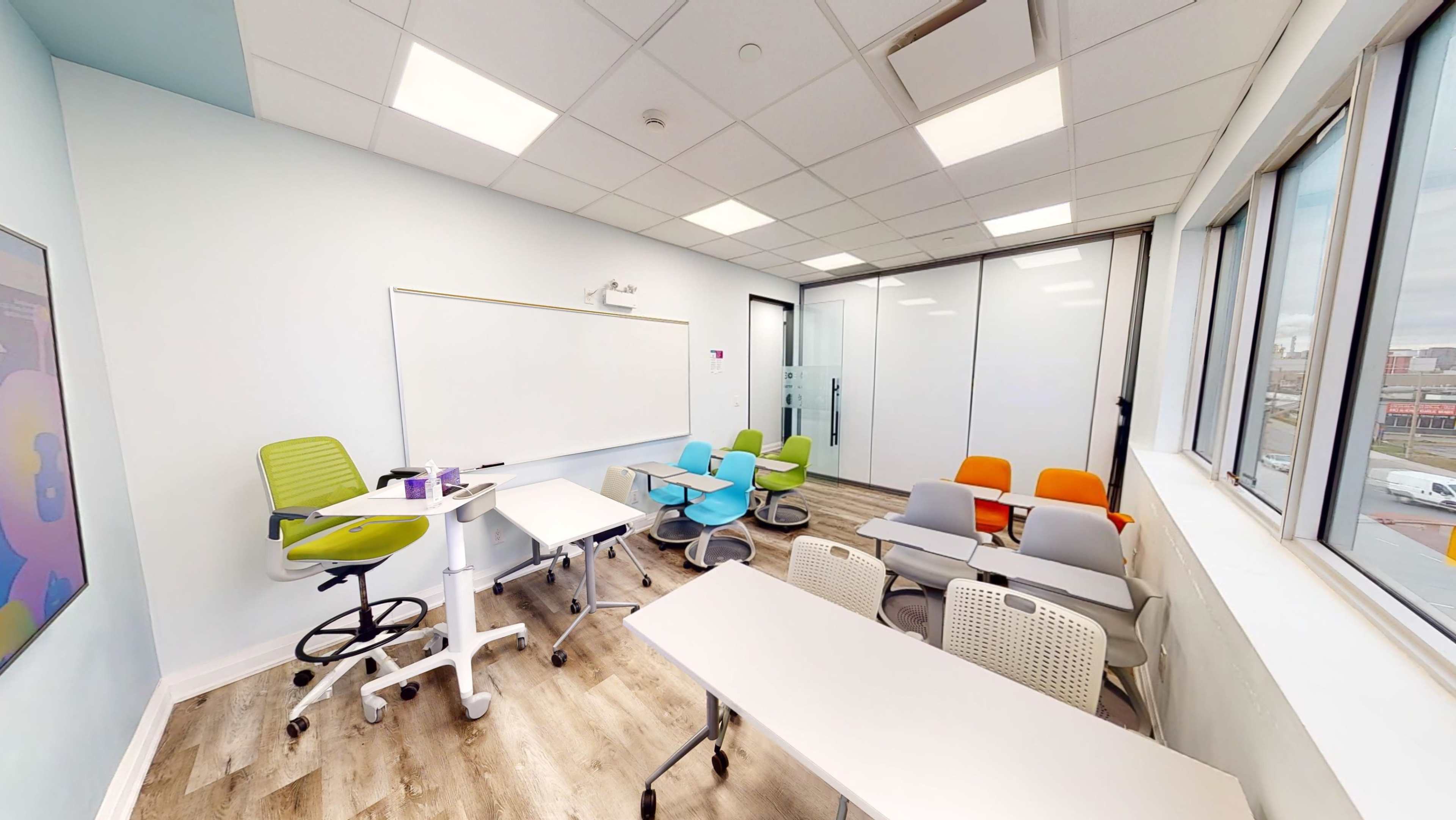 A modern classroom equipped with movable furniture, a whiteboard, and large windows offering natural light.