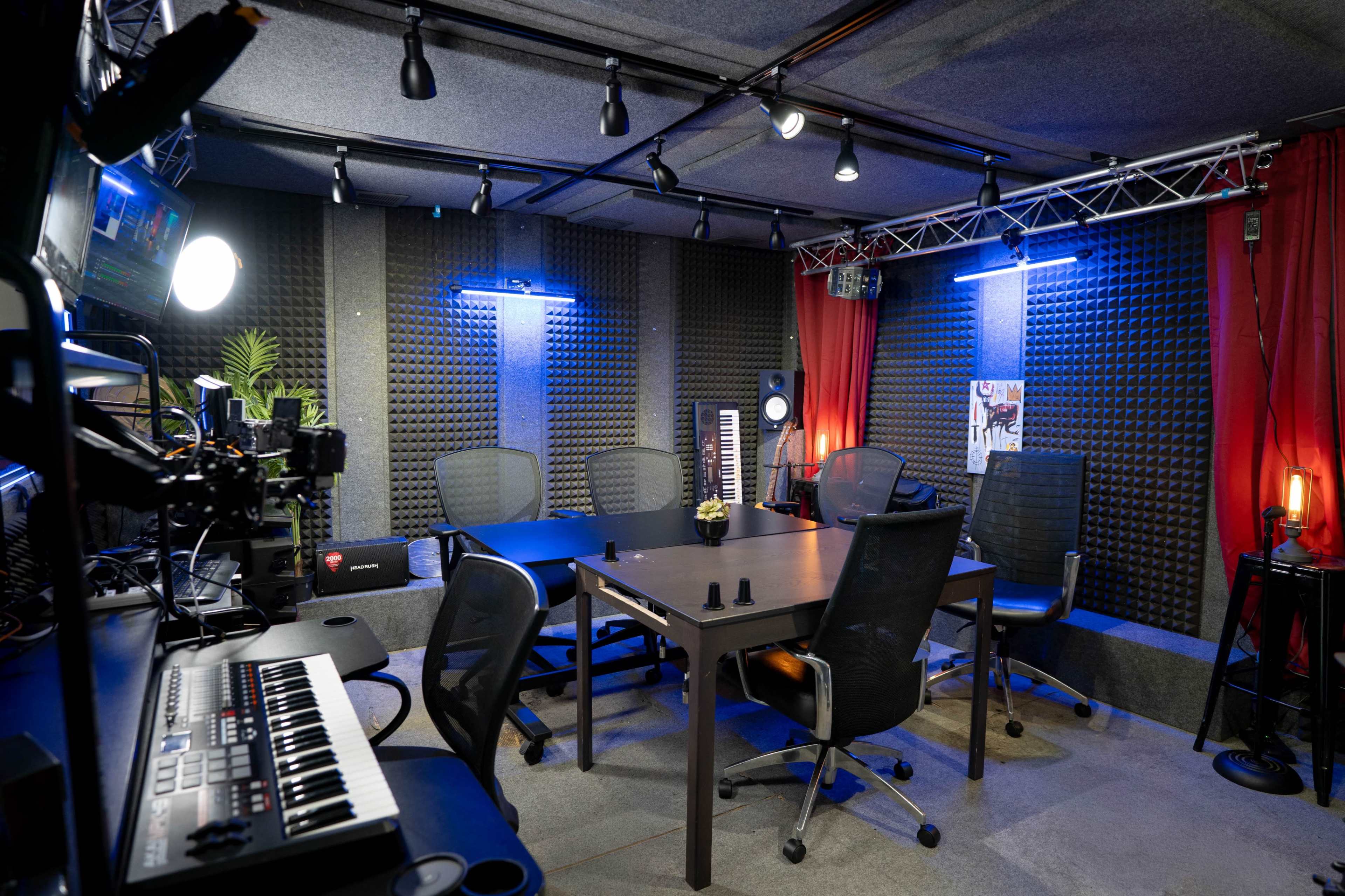 The image shows a music studio with soundproofed walls, a central table surrounded by chairs, and equipment including a keyboard and recording setup.
