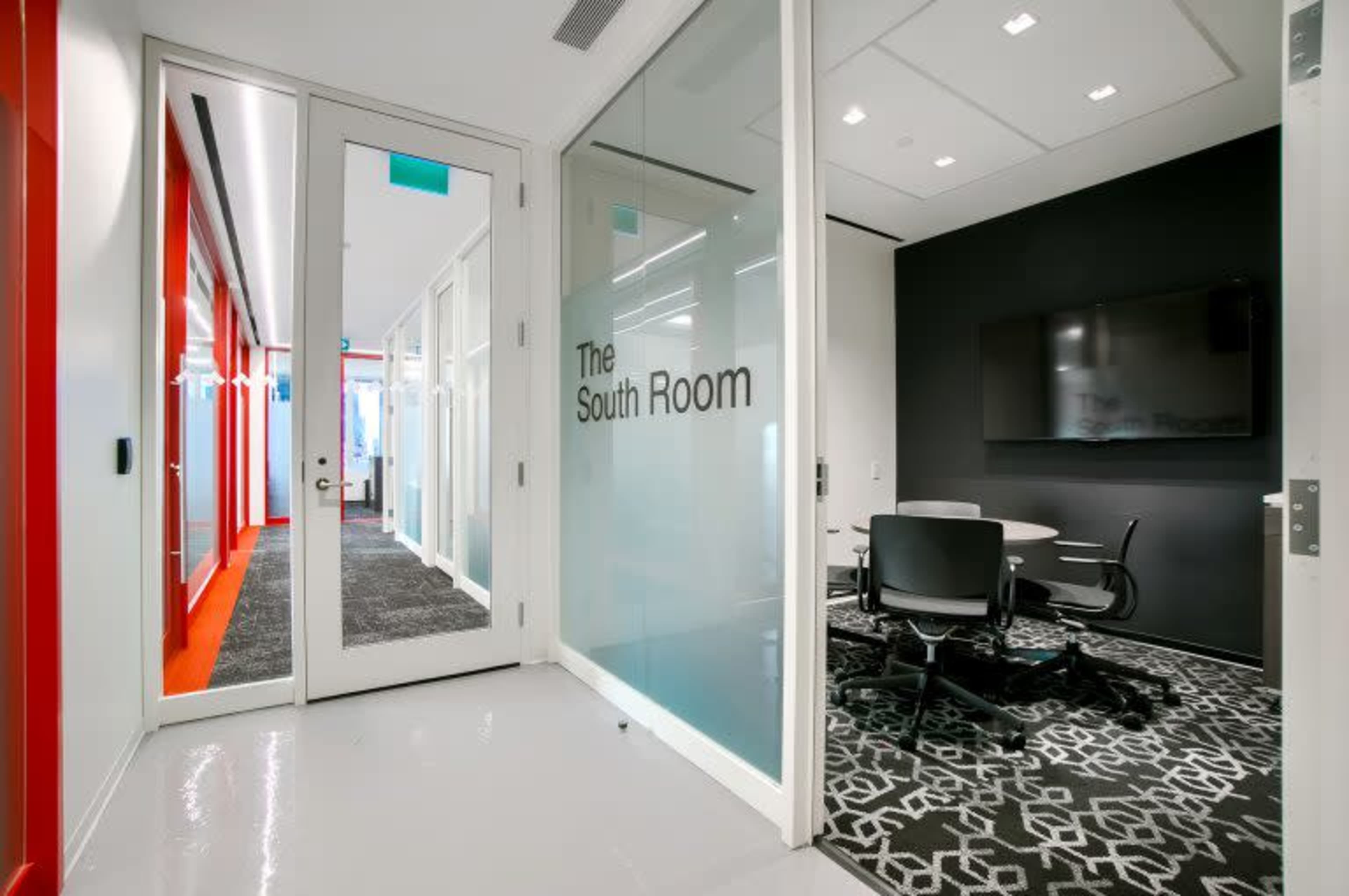 The image shows a modern office space with a glass-walled meeting room labeled "The South Room" and a dark wall featuring a wall-mounted screen.