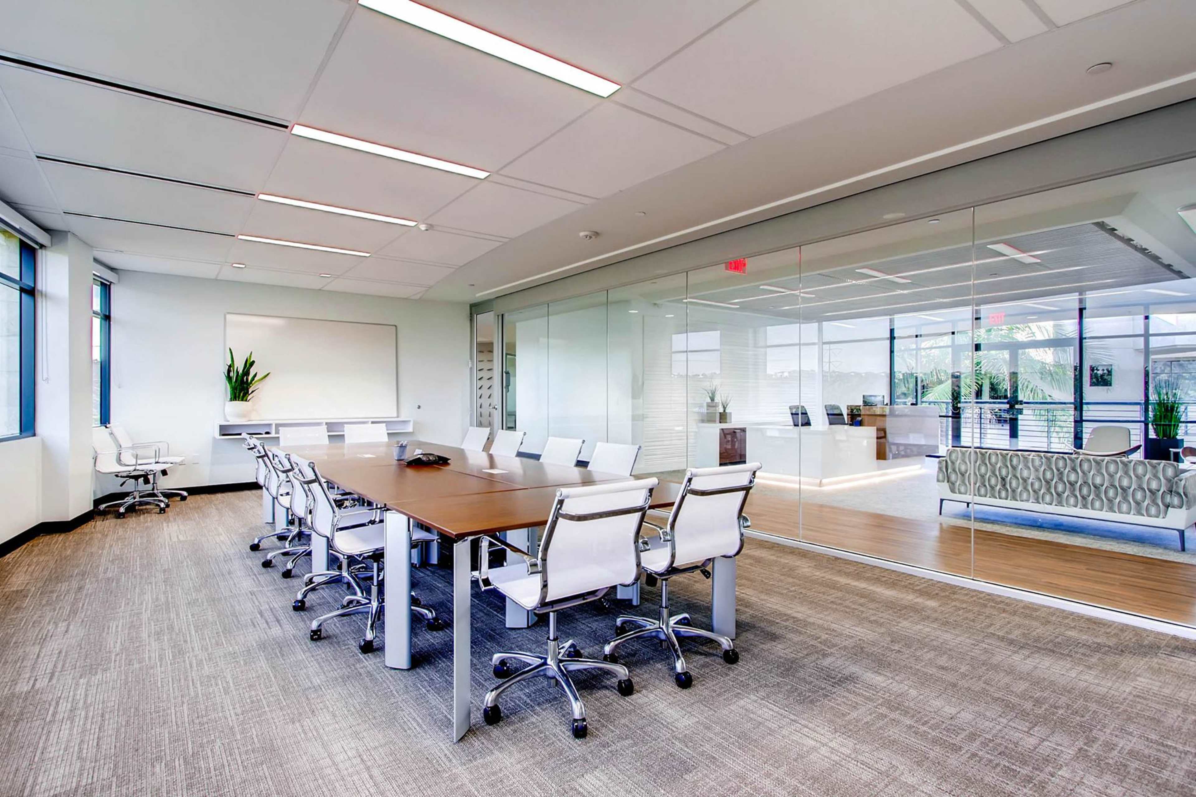 A large conference room features a long wooden table surrounded by white chairs, with glass walls and a view of an office space.