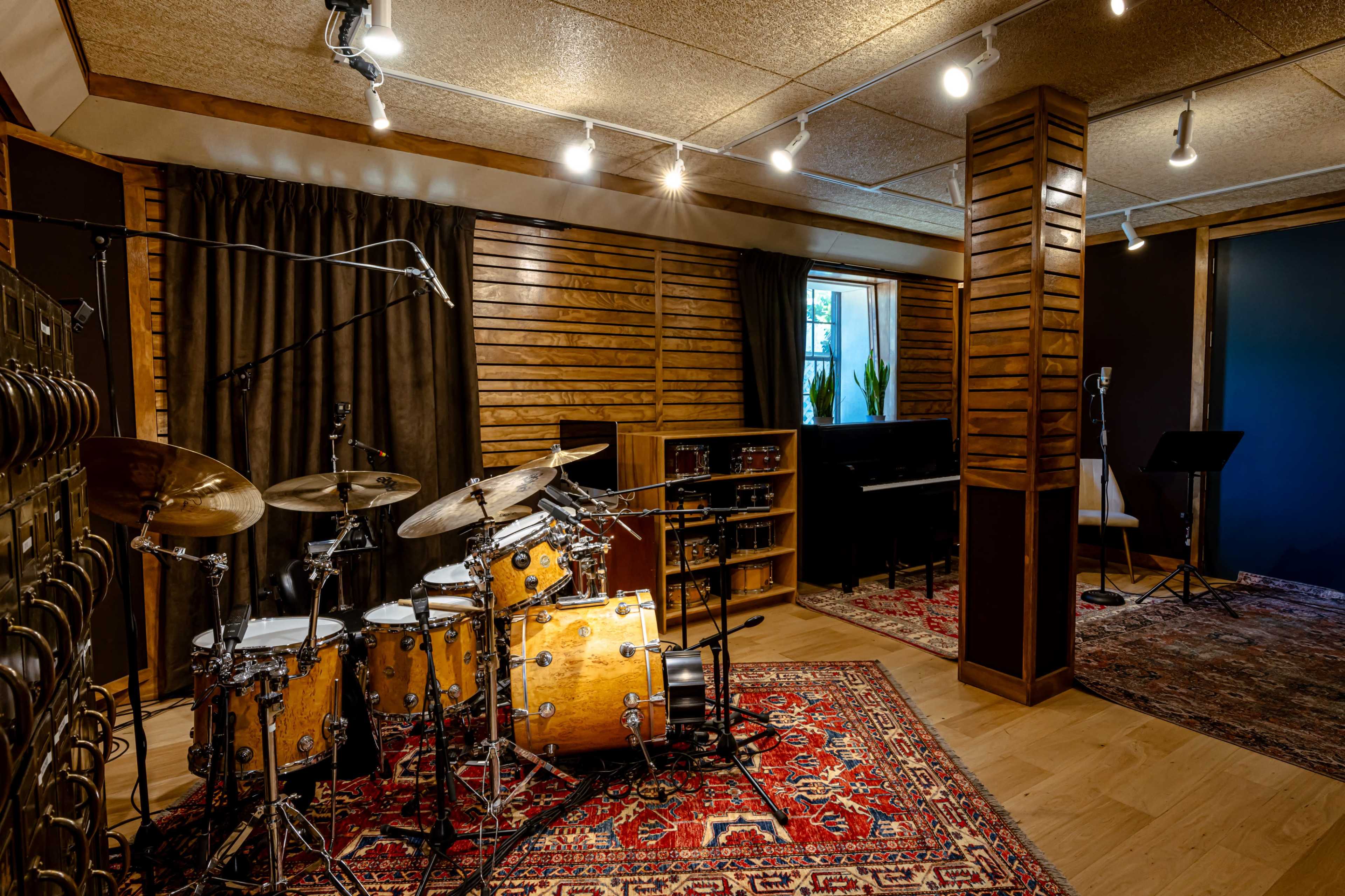 The image shows a professional music studio featuring a drum set, a piano, and soundproofed walls with warm wooden accents.
