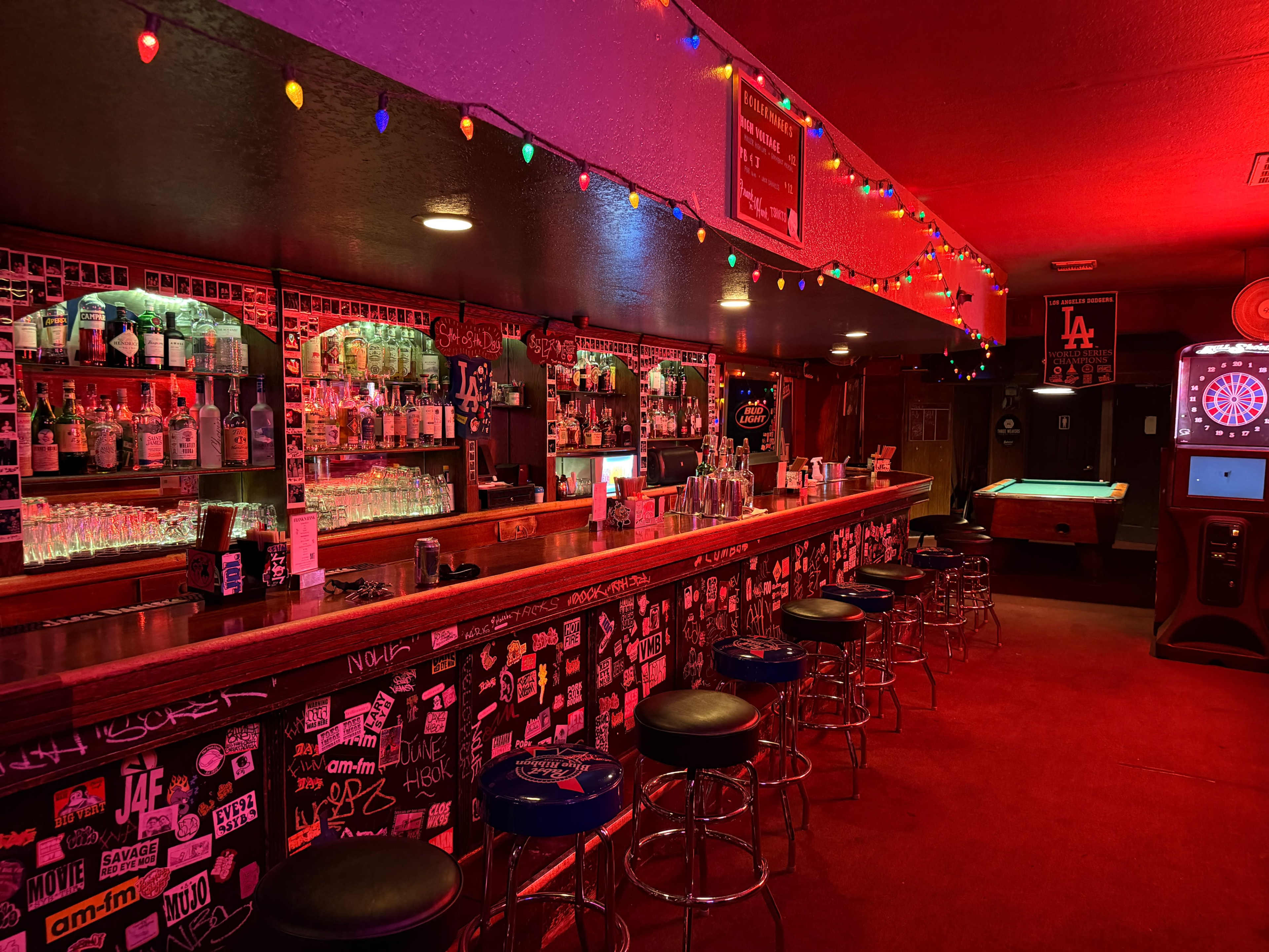 The image shows a dimly lit bar with a long counter, illuminated by colorful string lights, featuring shelves stocked with bottles and a pool table in the corner.