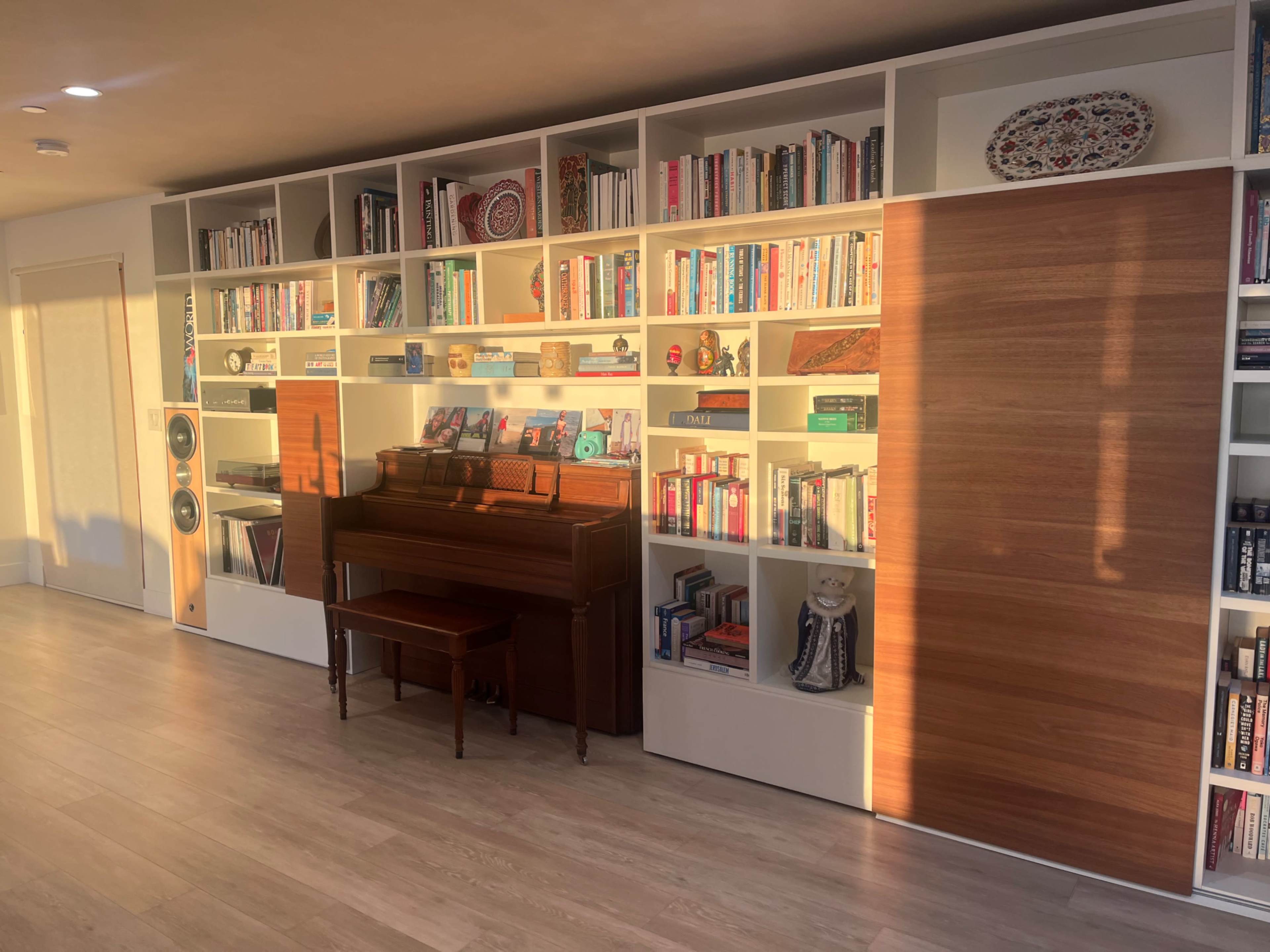 A wooden piano is positioned in front of a wall of bookshelves filled with various books and decorative items, with sunlight casting a warm glow on the scene.