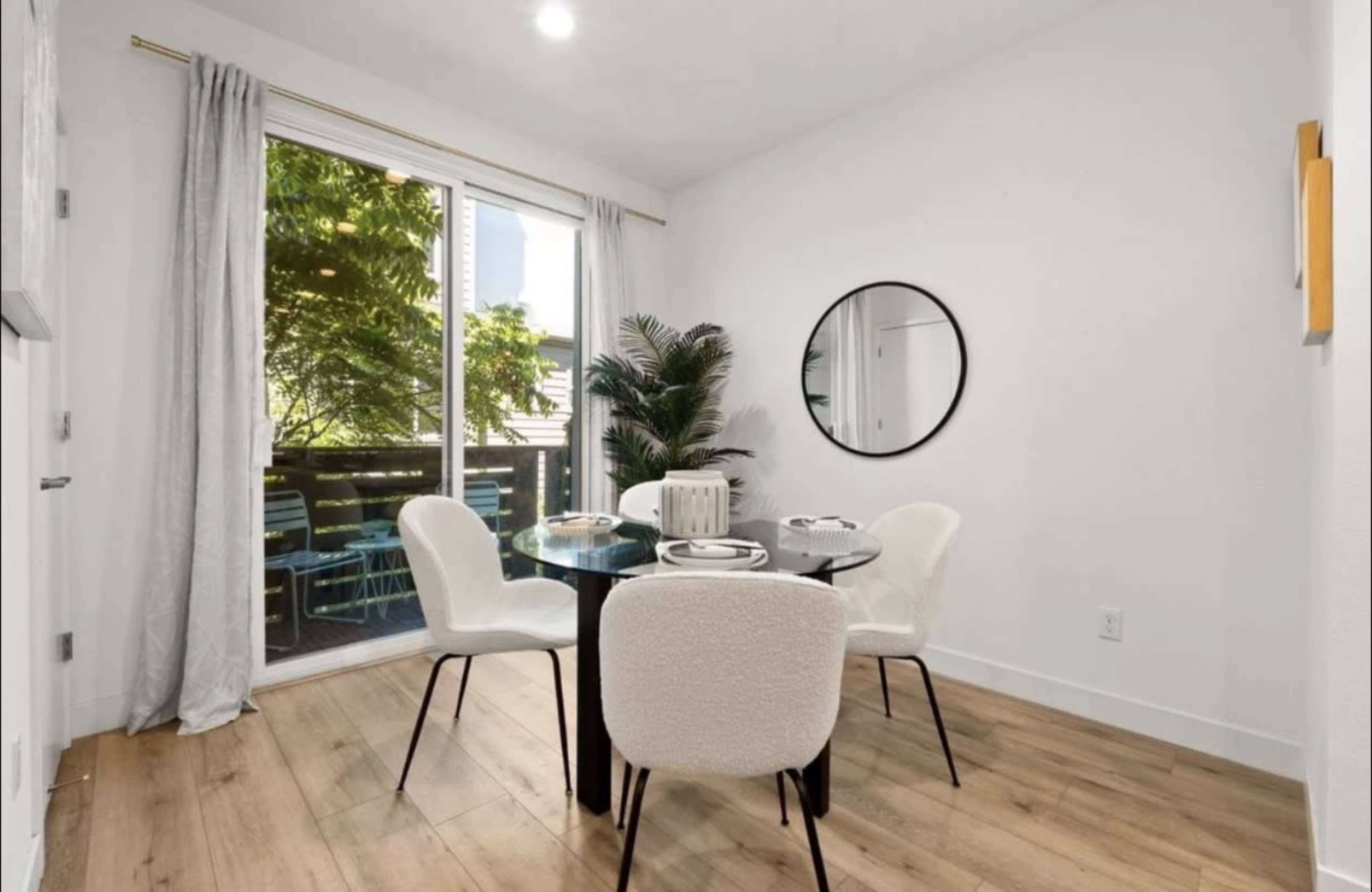 A dining area features a round glass table surrounded by four white chairs, with a large window providing a view of greenery outside.