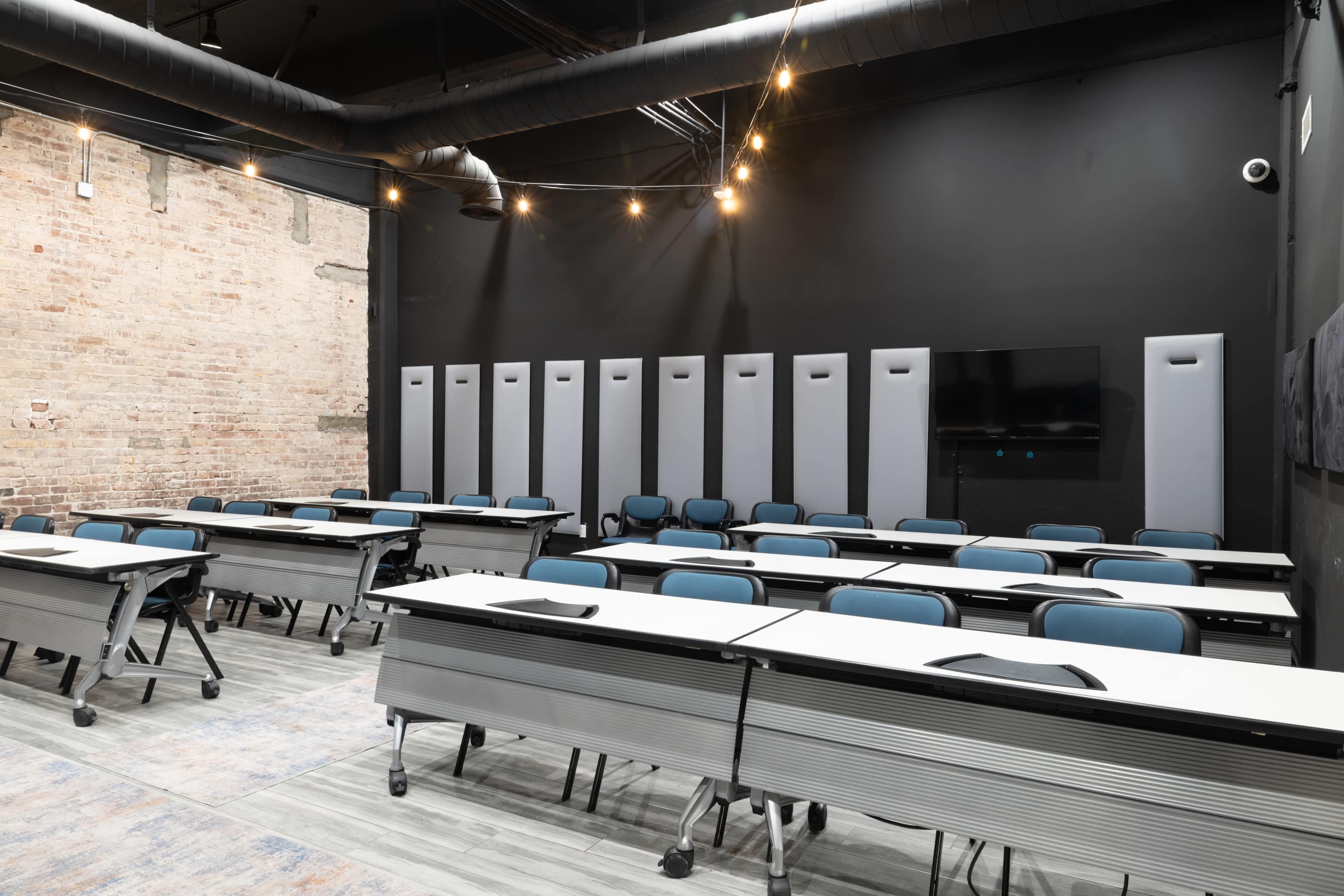 The image shows a modern classroom setup with rows of movable tables and chairs, against a backdrop of exposed brick and dark walls, illuminated by overhead lights.