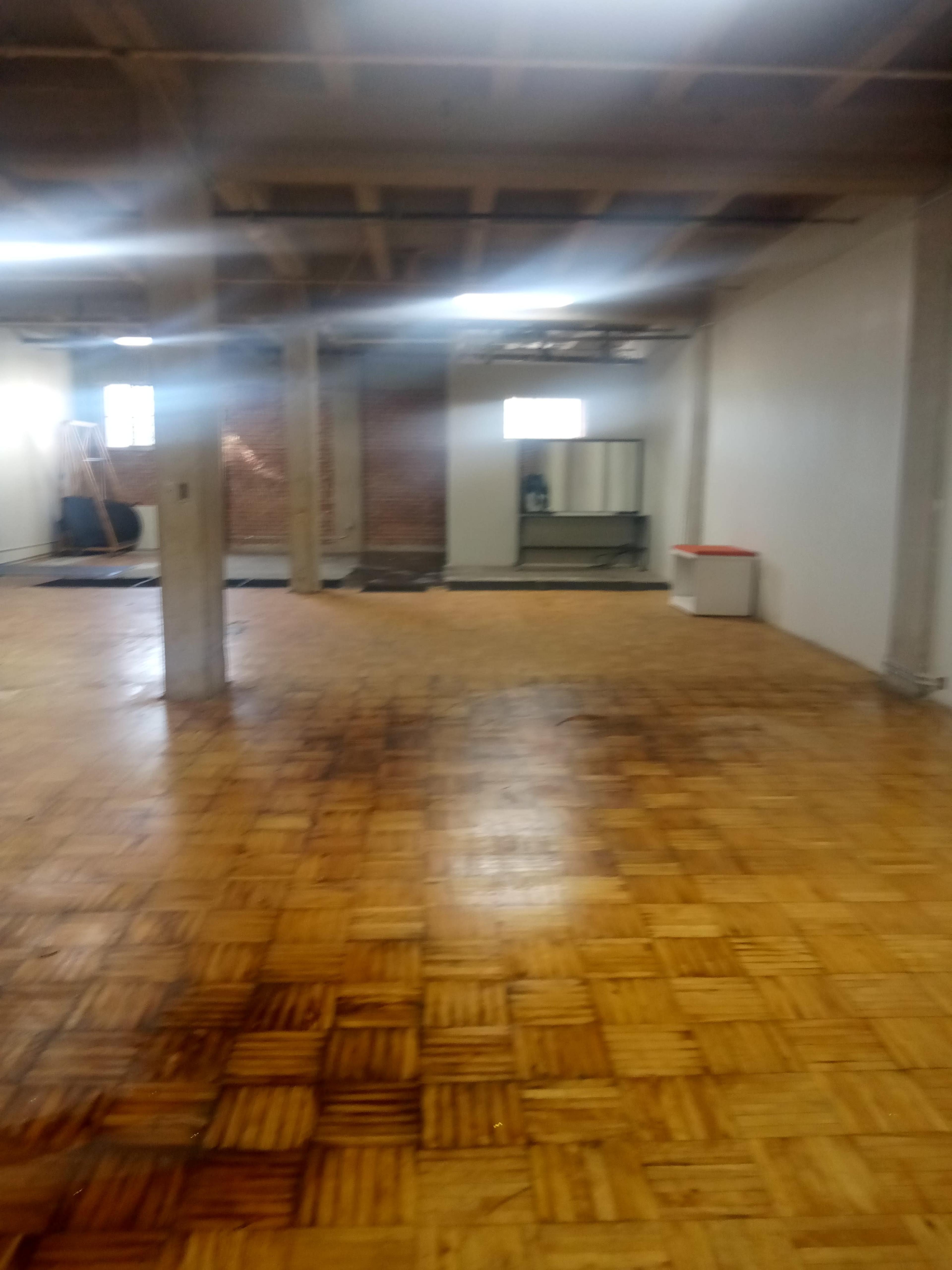 A large, empty room with wooden parquet flooring, exposed brick walls, and a few pieces of furniture in the background.