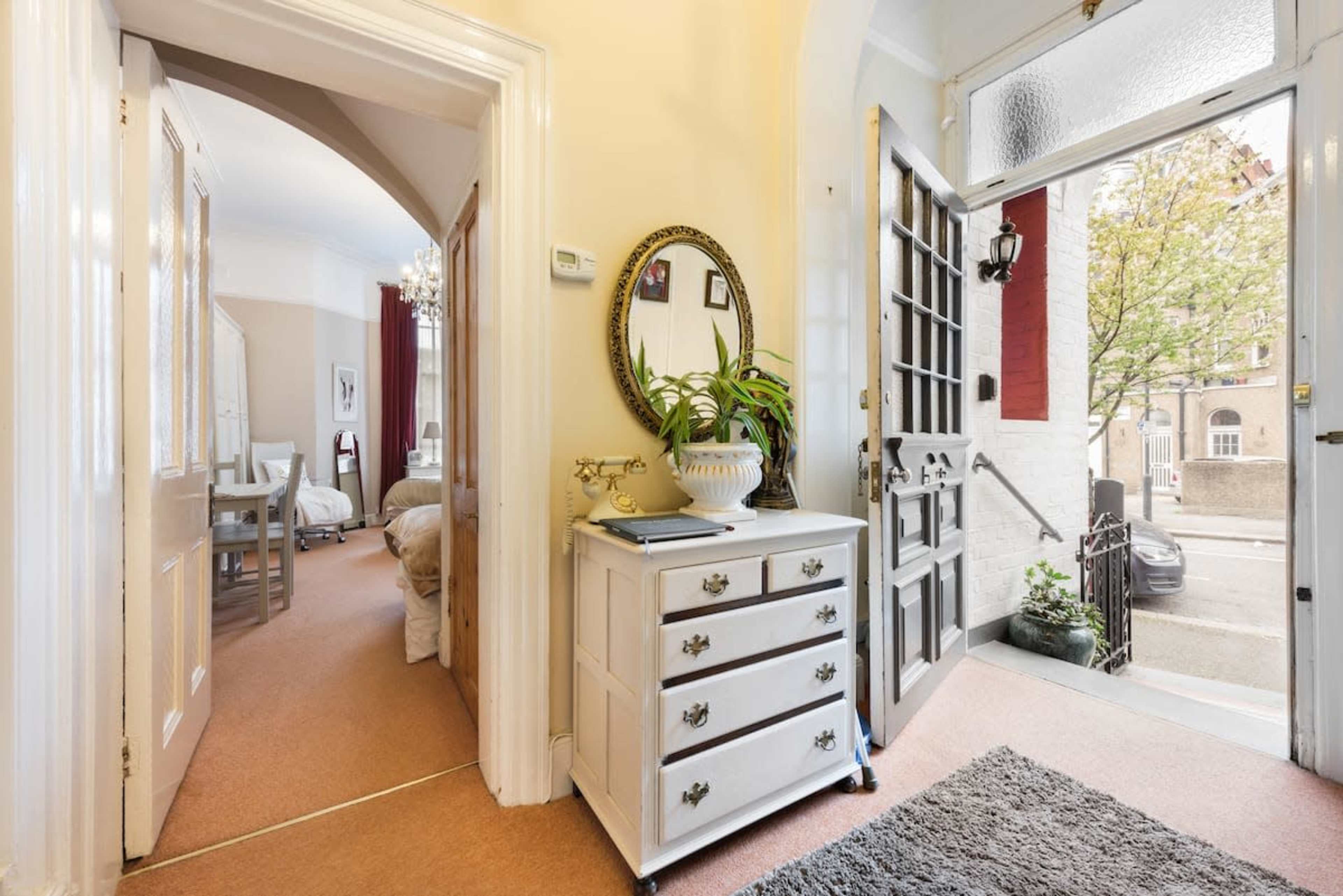 An entryway with a decorative mirror above a white chest of drawers, leading to an open door that reveals a view of the street outside.