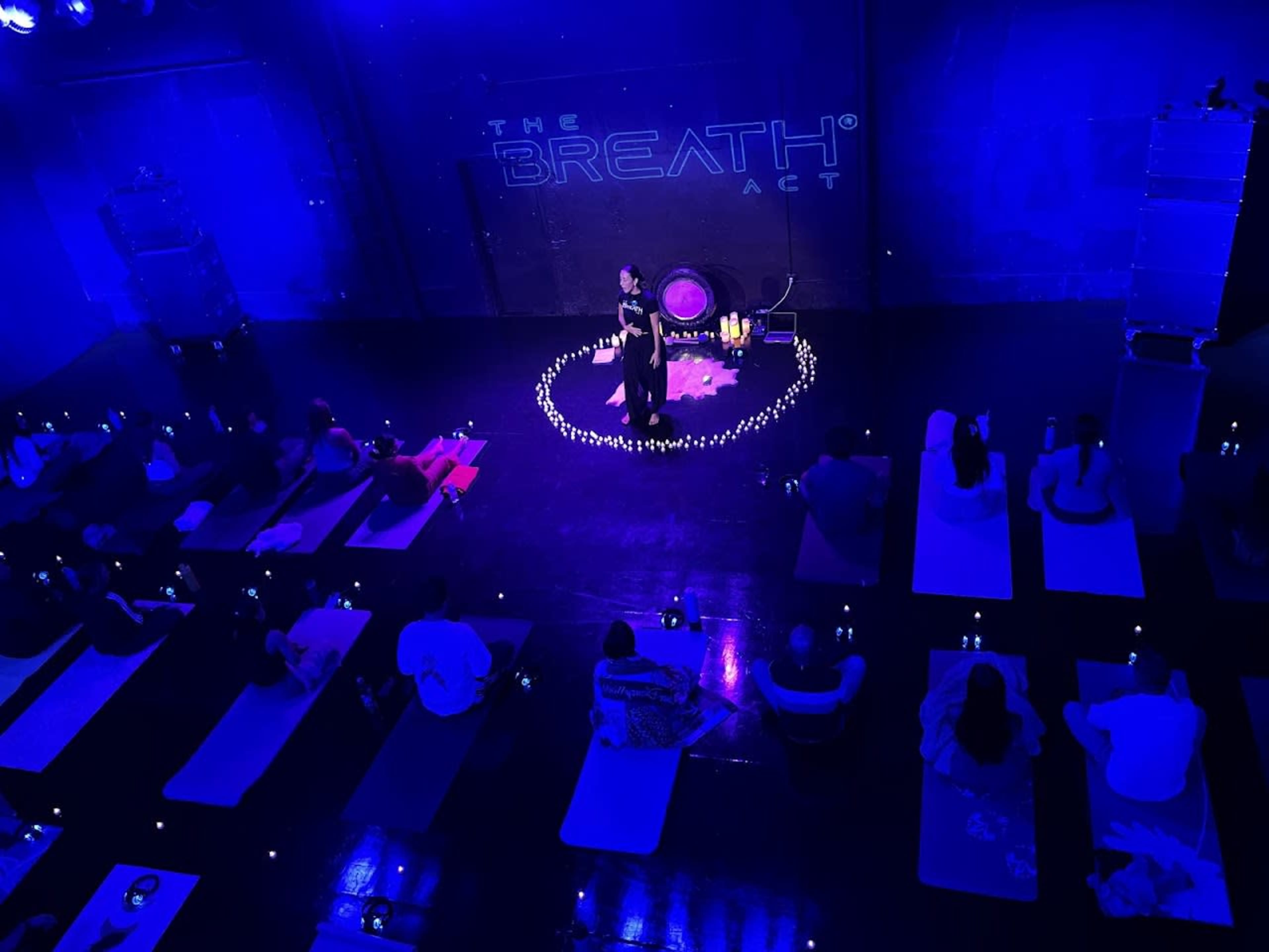 A group of people participates in a guided breathing session on yoga mats arranged in a circle, surrounded by candles, under dim blue lighting.
