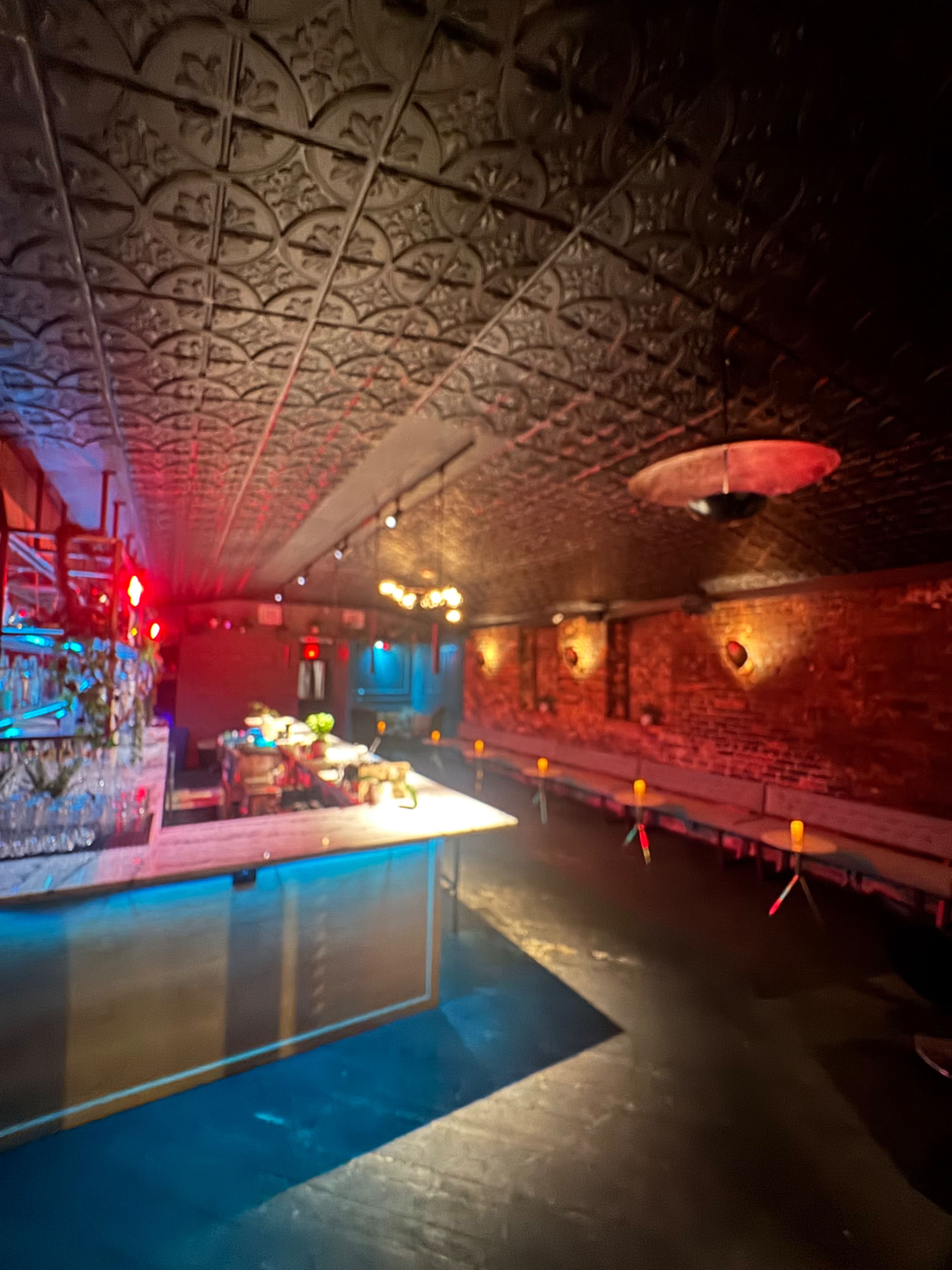 The image shows a dimly lit interior of a bar or lounge featuring a long bar countertop, decorative ceiling, and seating along the walls.