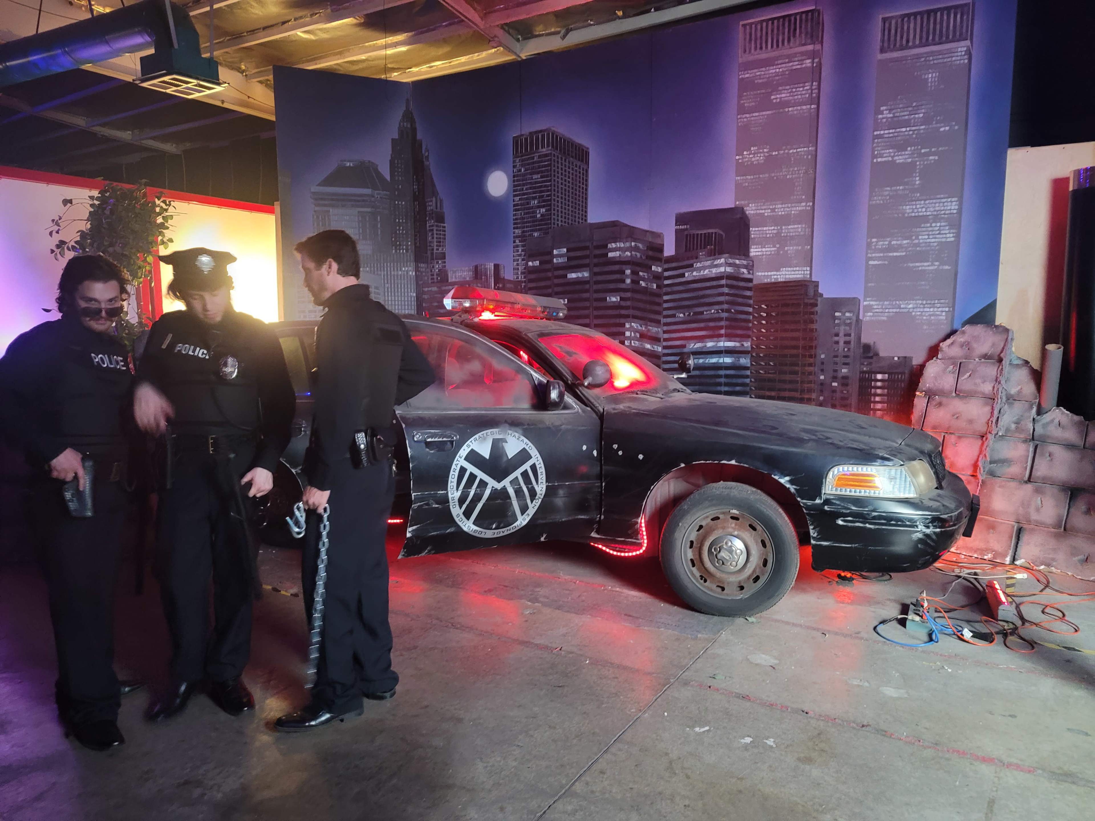 Two police officers stand beside a police car featuring a large emblem, set against a backdrop of a city skyline.