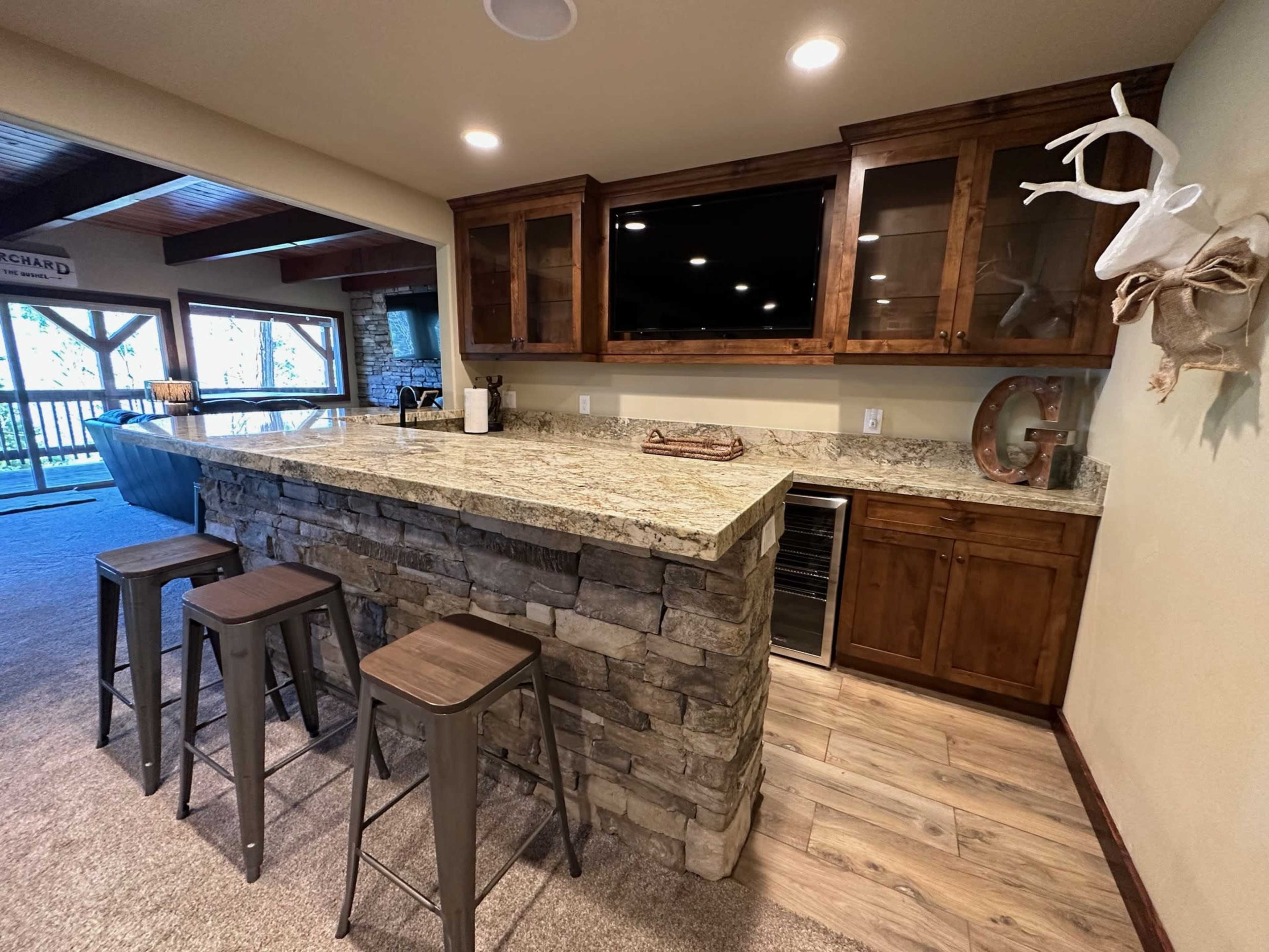 A kitchen bar area featuring a stone countertop, wooden cabinetry, a wall-mounted television, and metal stools.