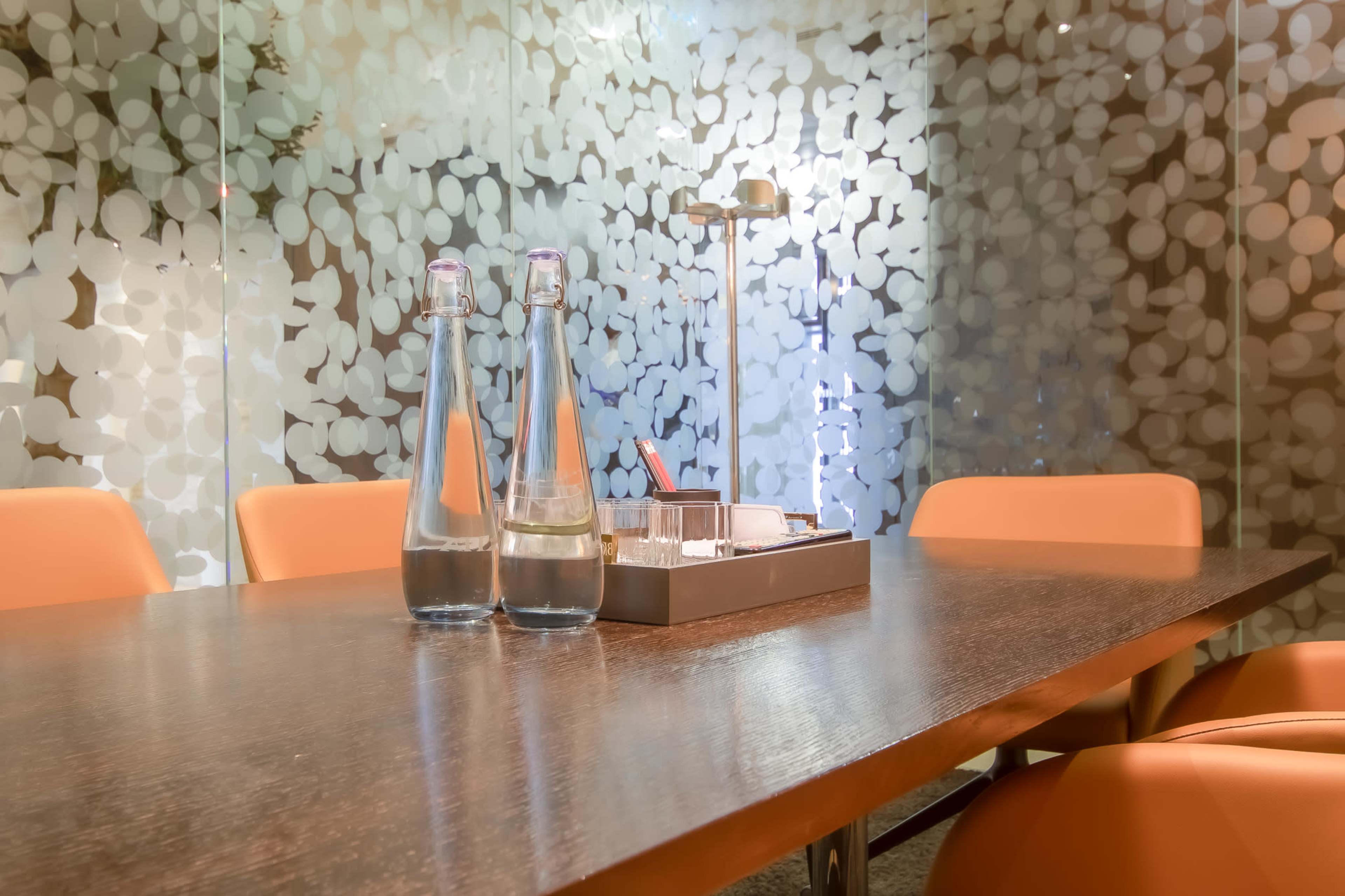 A conference room features a wooden table with two glass water bottles and a small tray on it, surrounded by orange chairs and a backdrop of frosted glass with a leaf pattern.