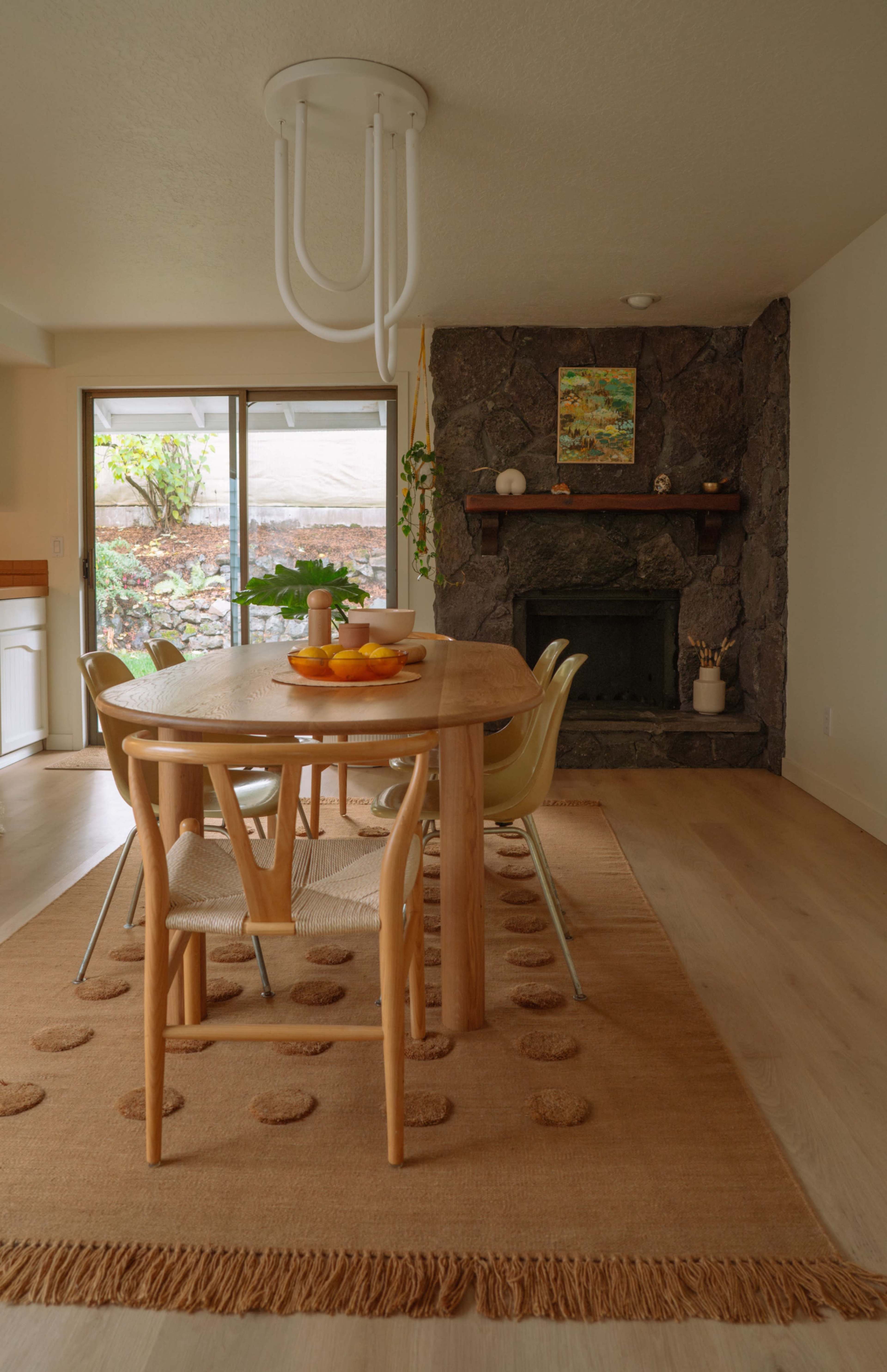 A wooden dining table with four chairs is set in a light-filled room, featuring a stone fireplace and large windows.