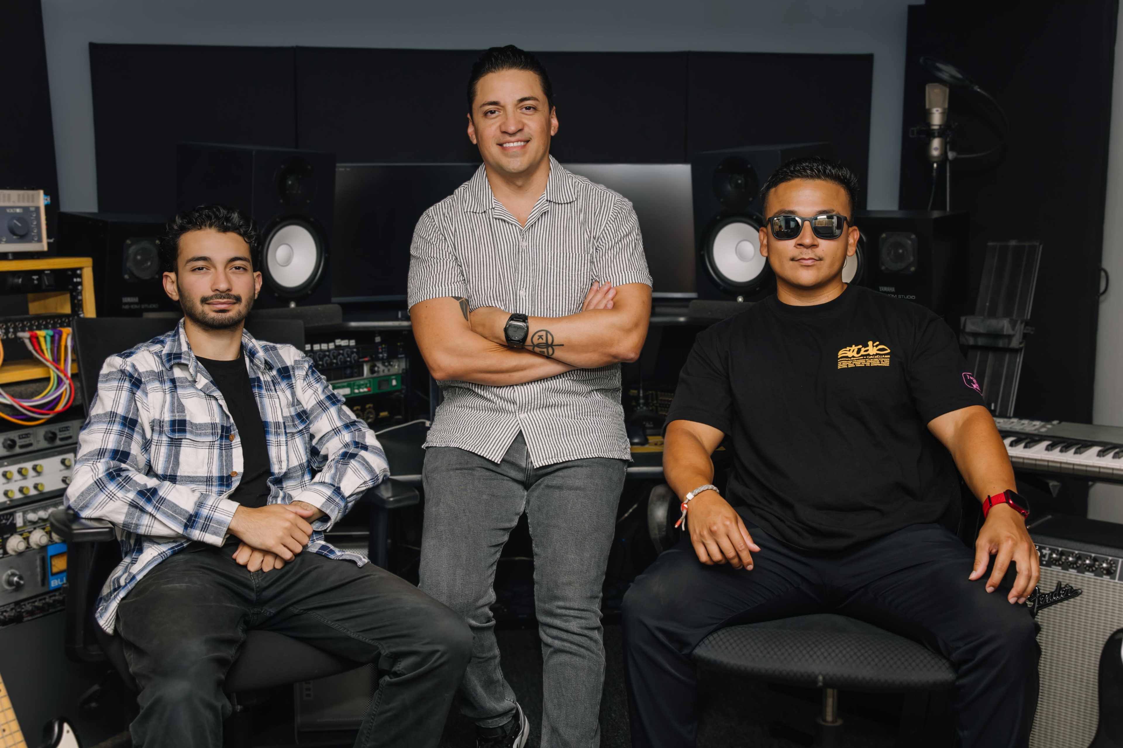 Three men are seated in a music studio surrounded by audio equipment and speakers.